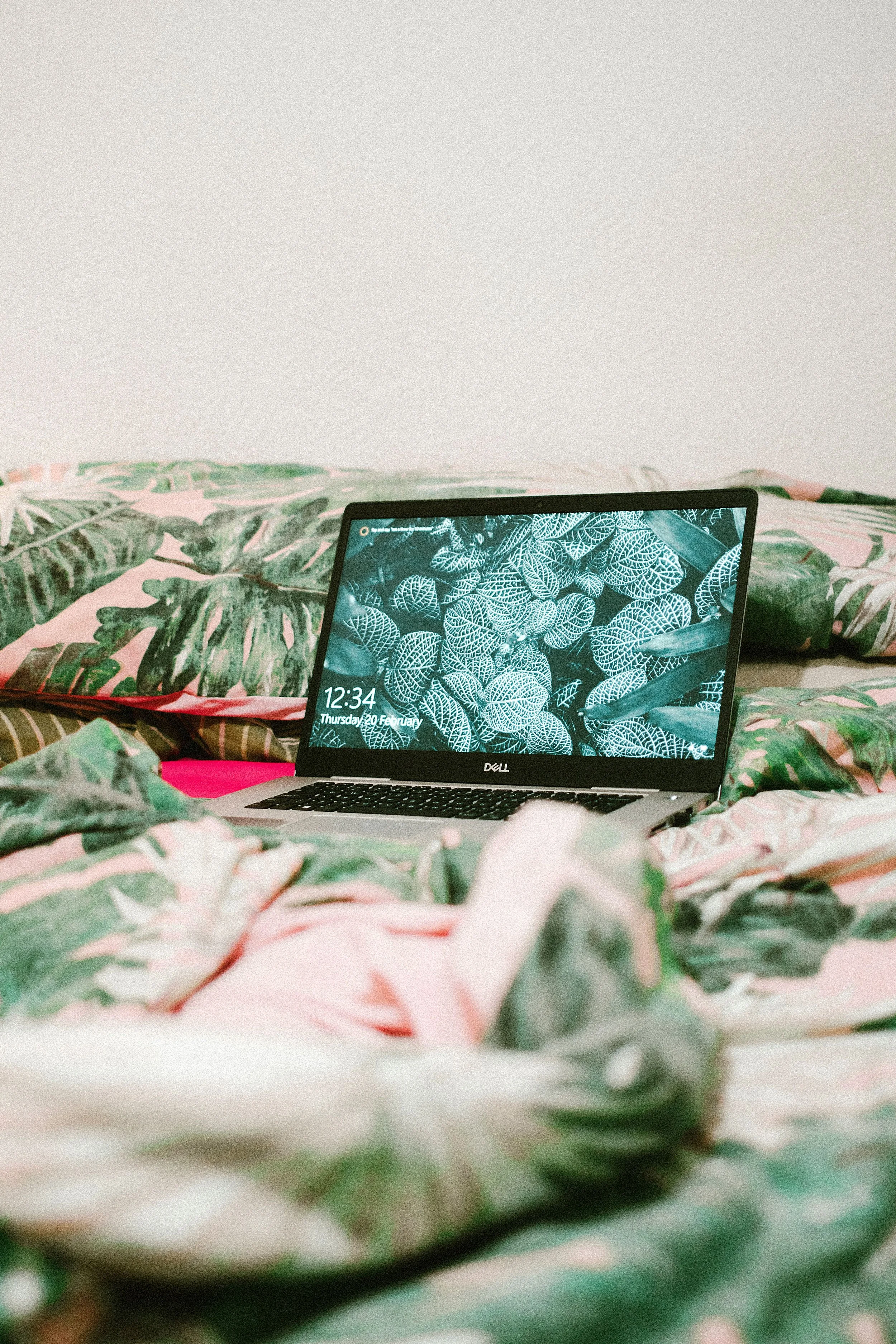 A laptop on a bed with tropical leaf patterned bedding, displaying the time 12:34 on February 20th.