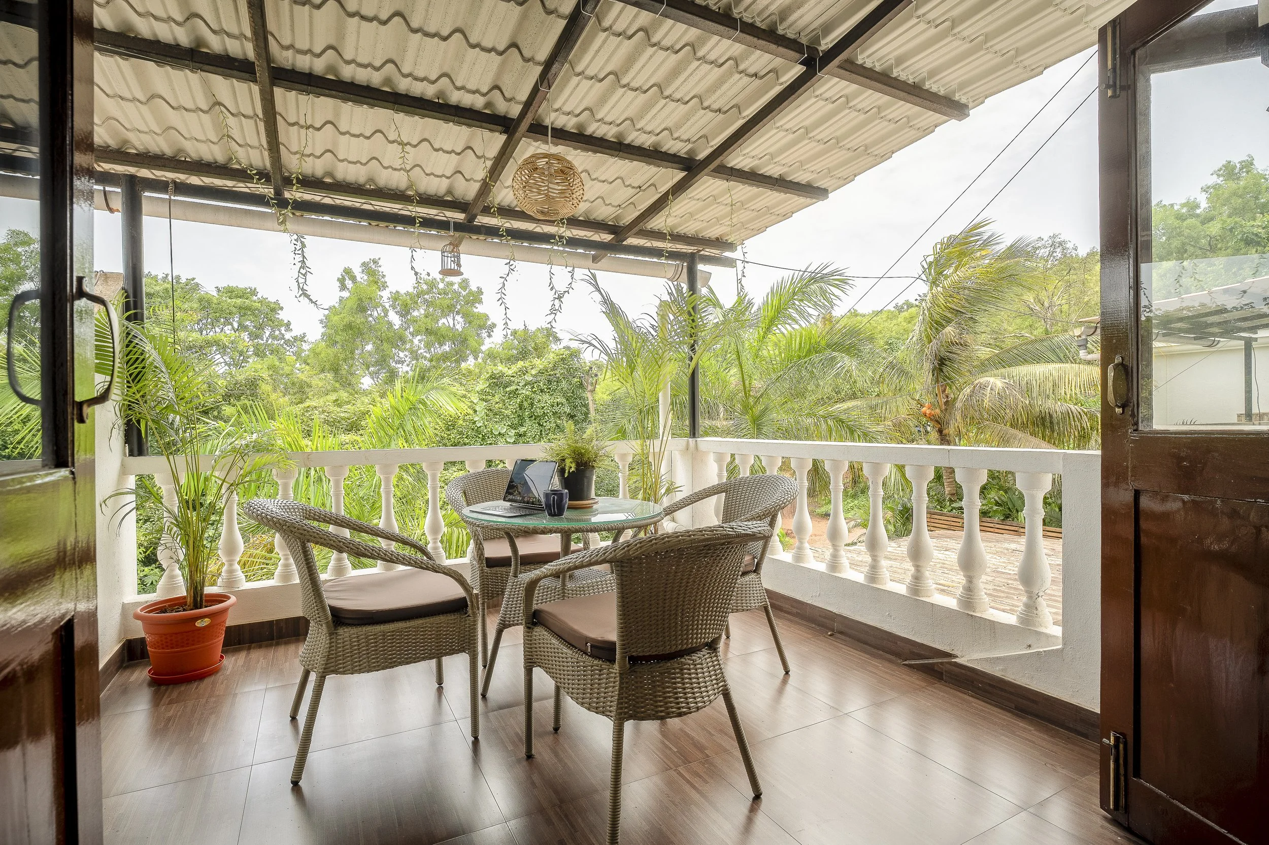 3BHK Villa In Goa With A covered outdoor patio with a round table, four wicker chairs, potted plants, and a view of lush green trees and sky.