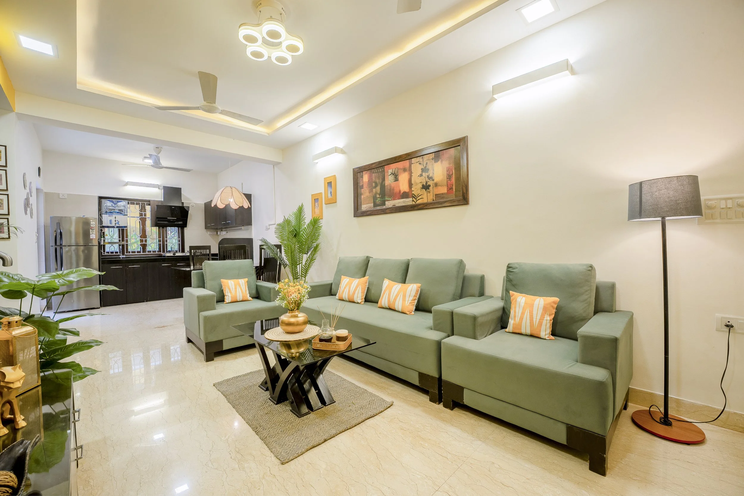 3BHK Villa In Goa With a Living room with green sofa, black coffee table with a floral arrangement, floor lamp, wall art, and kitchen in the background with a refrigerator and cabinets.