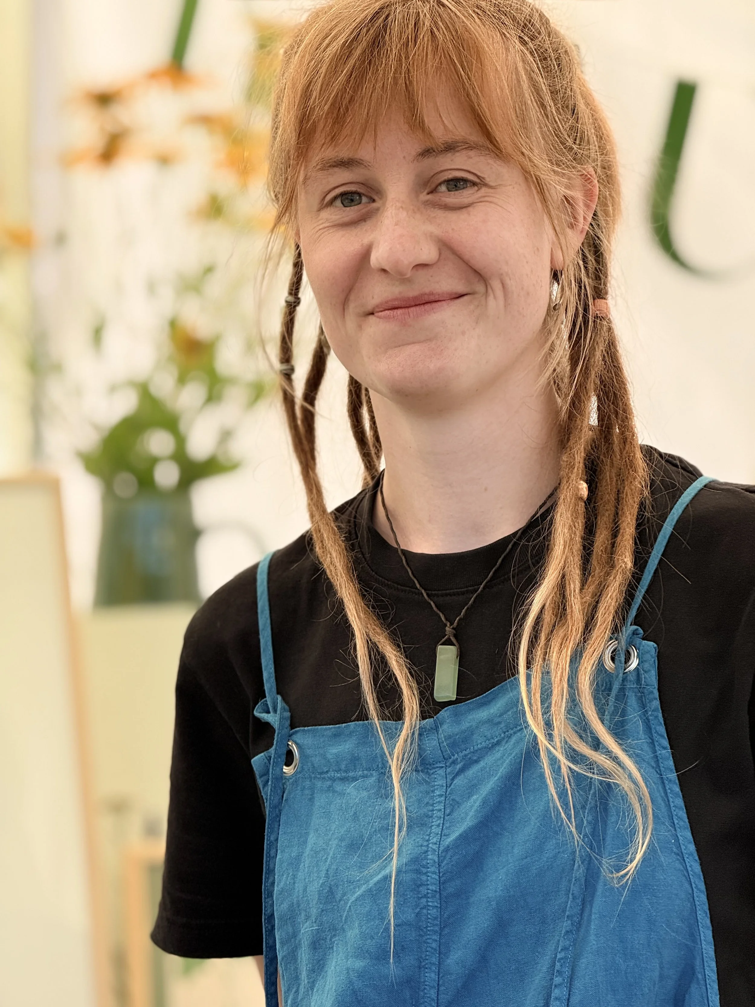 Eine lachende Frau mit roten Haaren, die in Dreadlocks geflochten ist, trägt einschwarzes T-Shirt und eine blaue Latzhose. Im Hintergrund sind Blumen und grüne Pflanzen sichtbar.