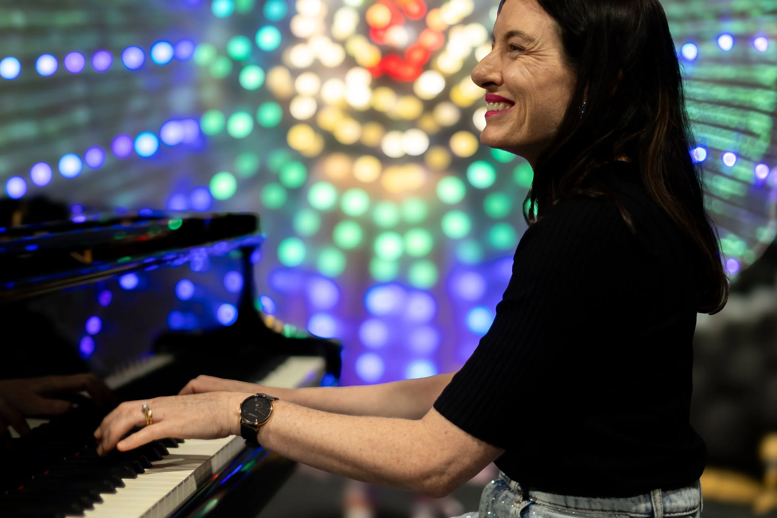A woman with dark hair and a smile playing the piano in an environment with colorful, blurred light decorations in the background.