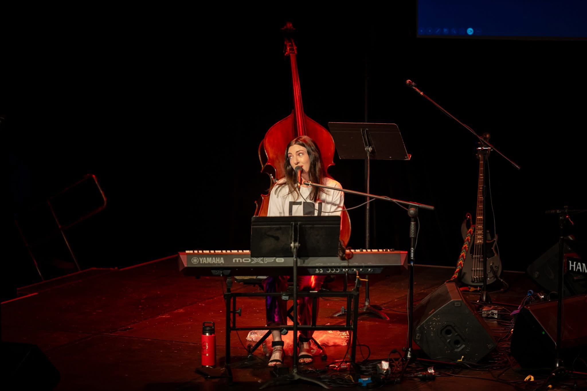 A woman performing on stage with a keyboard and microphone. There is a double bass and electric bass in the background, in a dimly lit setting.