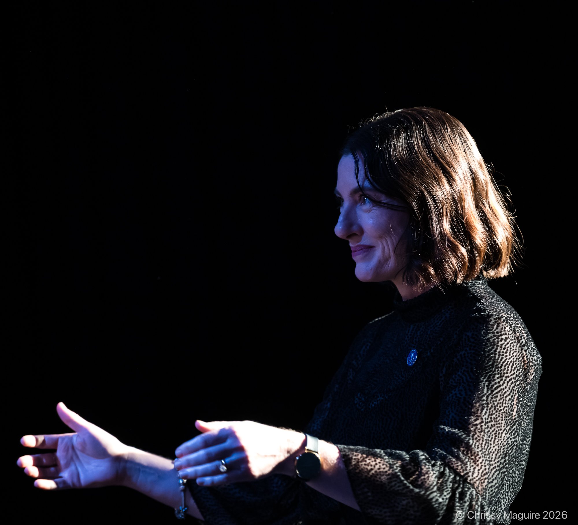 A woman with wavy, light brown hair is looking at a choir, smiling. She is wearing a black top and has jewellery, including rings and a bracelet. Her hands are out in front of her, ready to conduct the choir.