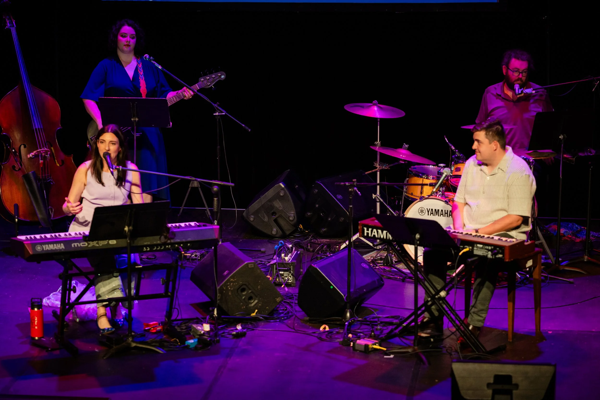 Musicians performing on stage with various instruments including piano, bass, drums, and guitars, with stage lights illuminating them against a dark background.