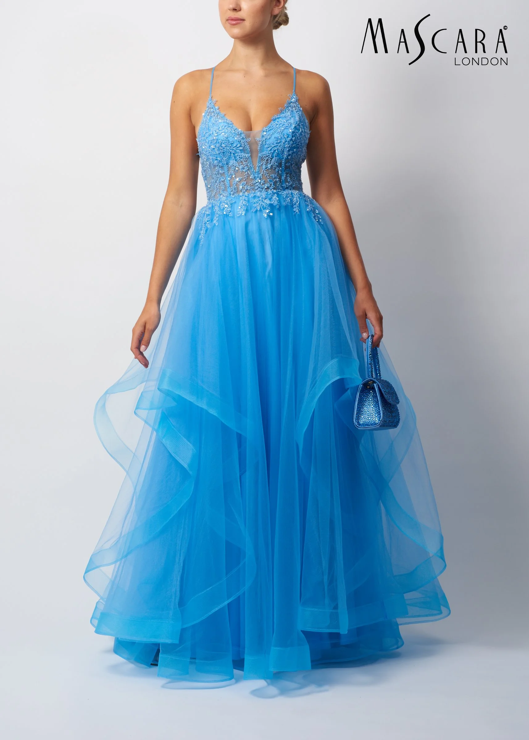 A woman in a blue evening gown with lace and beaded embroidery, holding a matching blue purse, standing against a plain white background with the Mascara London logo.