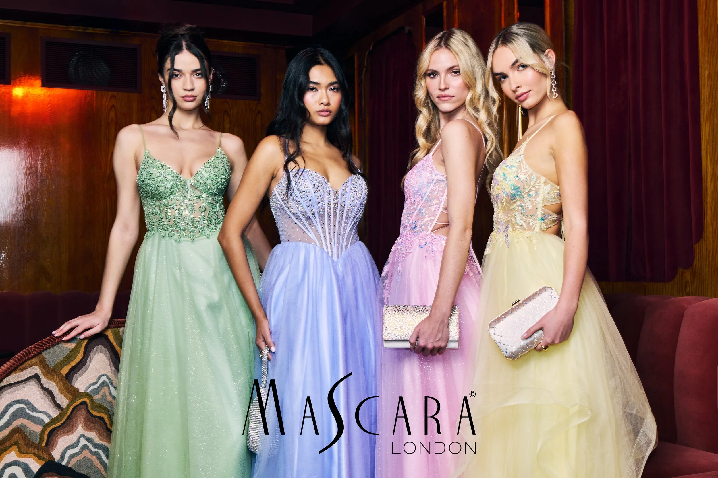 Four women in formal dresses standing indoors, with a partial logo reading "MASCARA London" visible in the foreground.