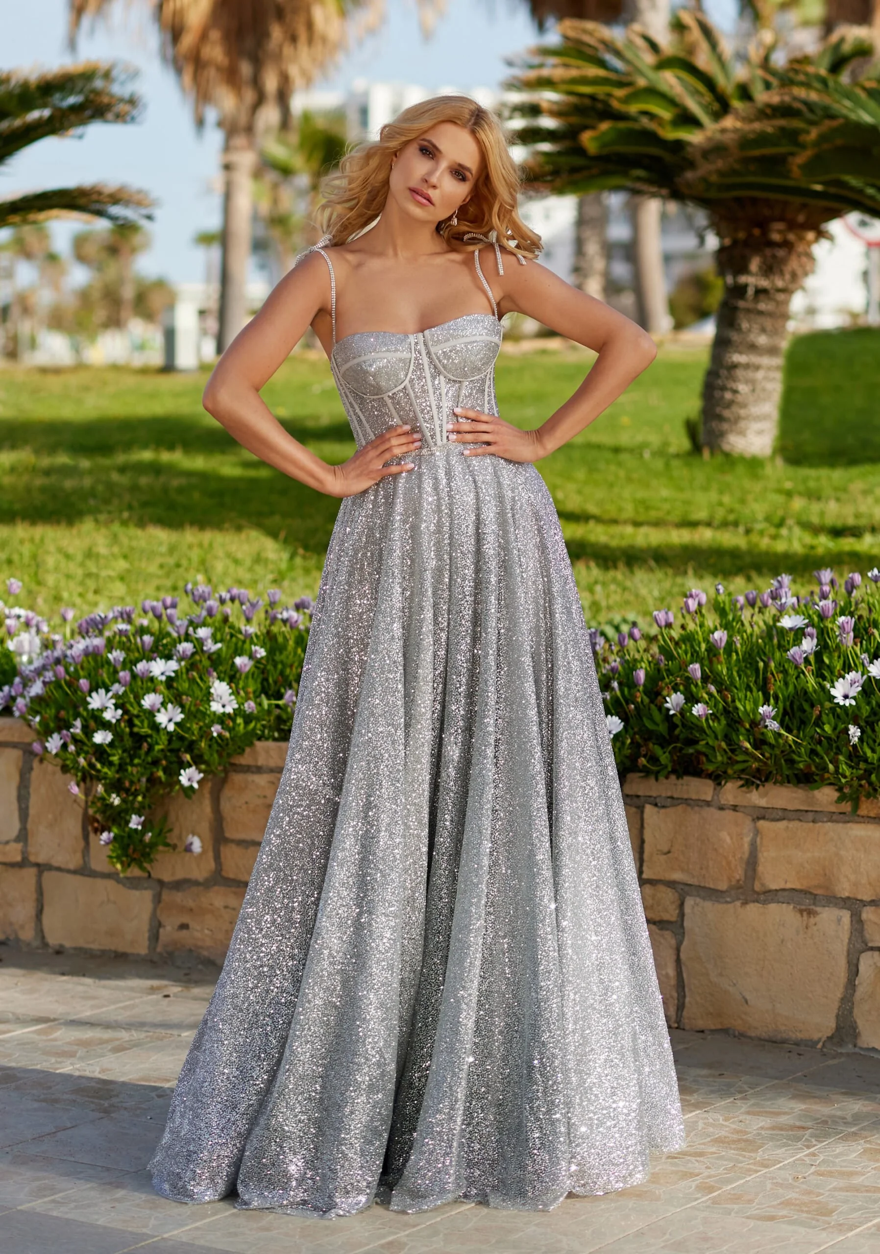 A woman in a shiny silver evening gown standing outdoors near a flower bed and palm trees, with a blurred cityscape in the background.