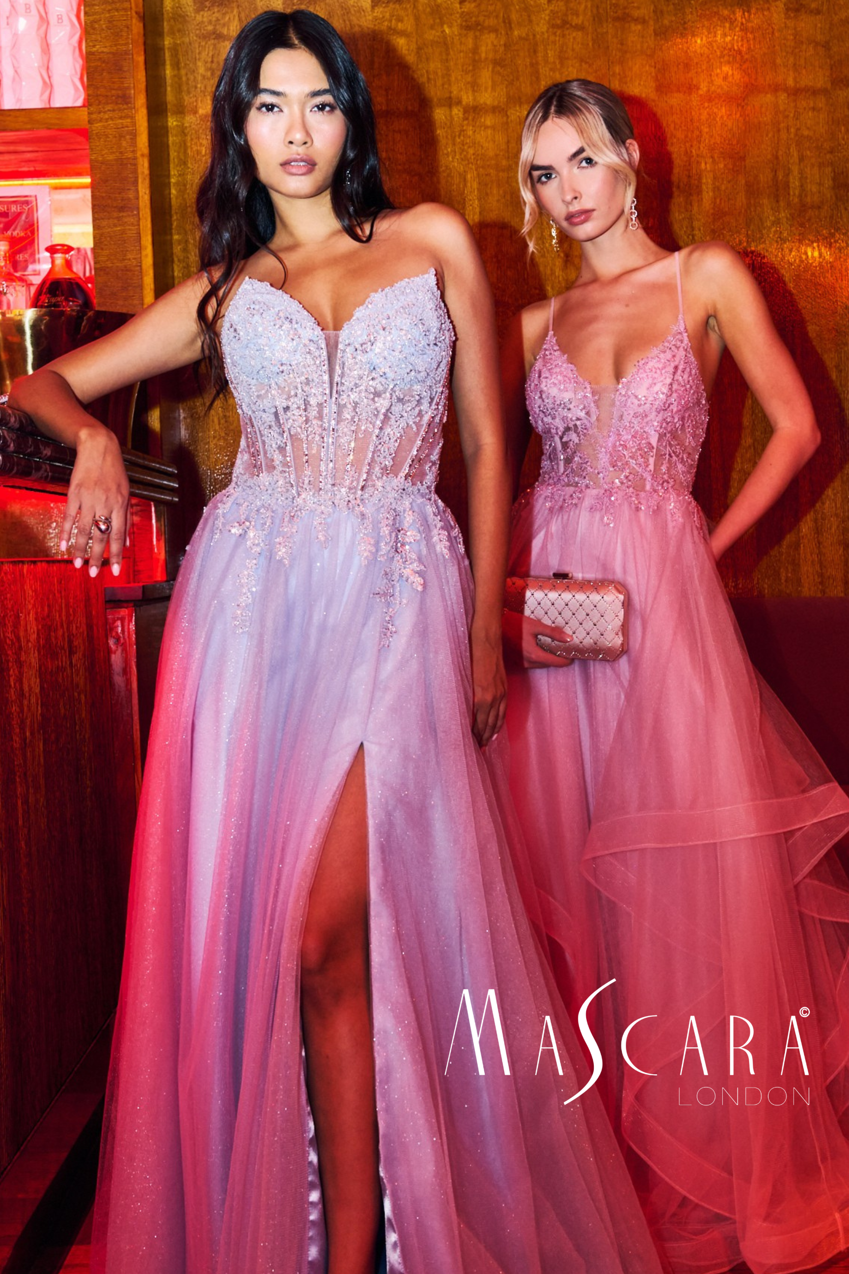 Two women in pink prom dresses standing against a wooden wall, one with black hair and the other with light brown hair, both posing confidently at an event with the words 'Mascara London' visible.