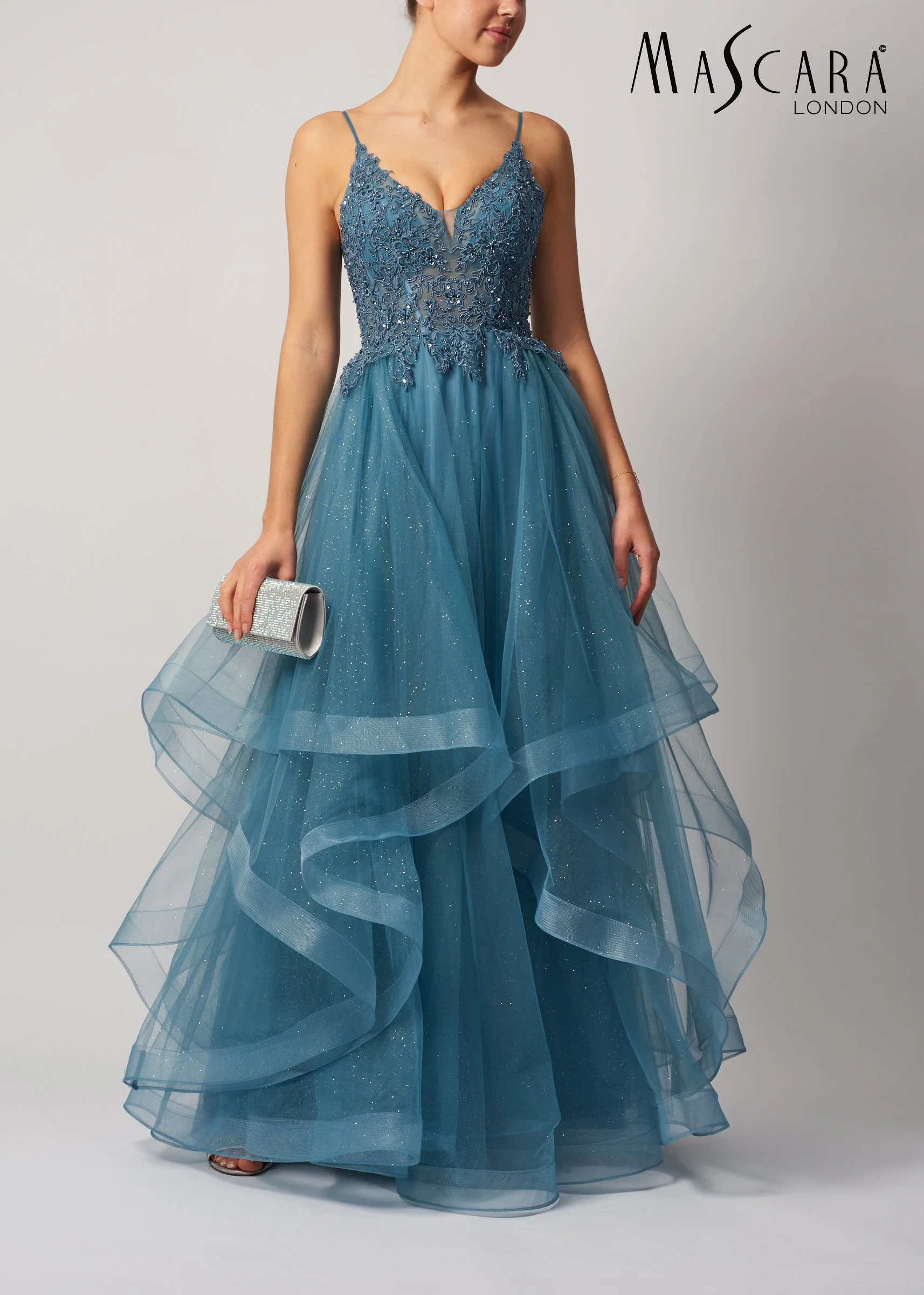 A woman in a strapless blue formal dress with intricate embroidery and a full tulle skirt, holding a silver clutch, standing against a plain background, with the text 'Mascara London' in the top right corner.