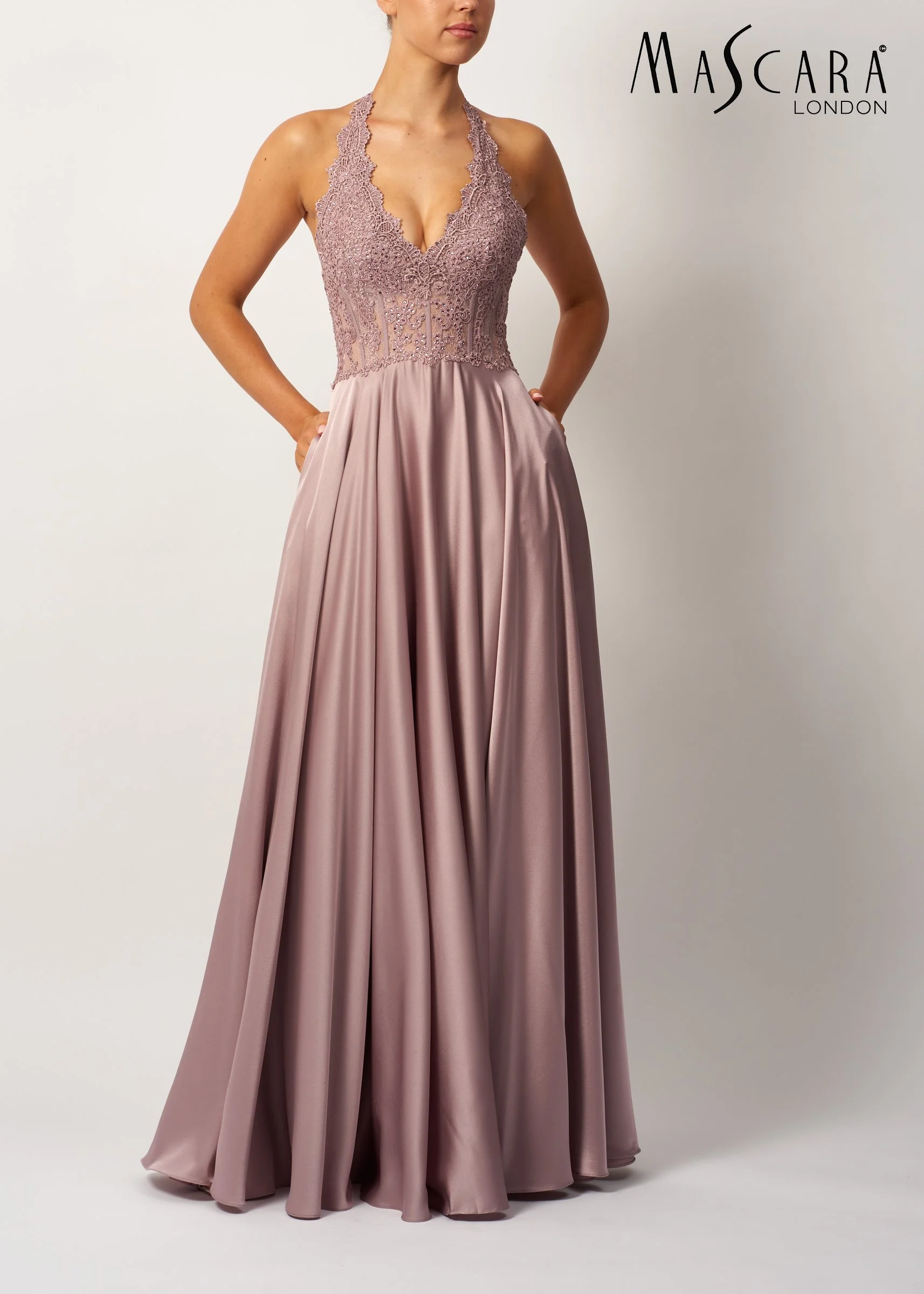 A woman wearing a sleeveless, lace and beaded mauve evening gown with a deep V-neck and pleated satin skirt, standing against a plain white background with the "Mascara London" logo in the top right corner.