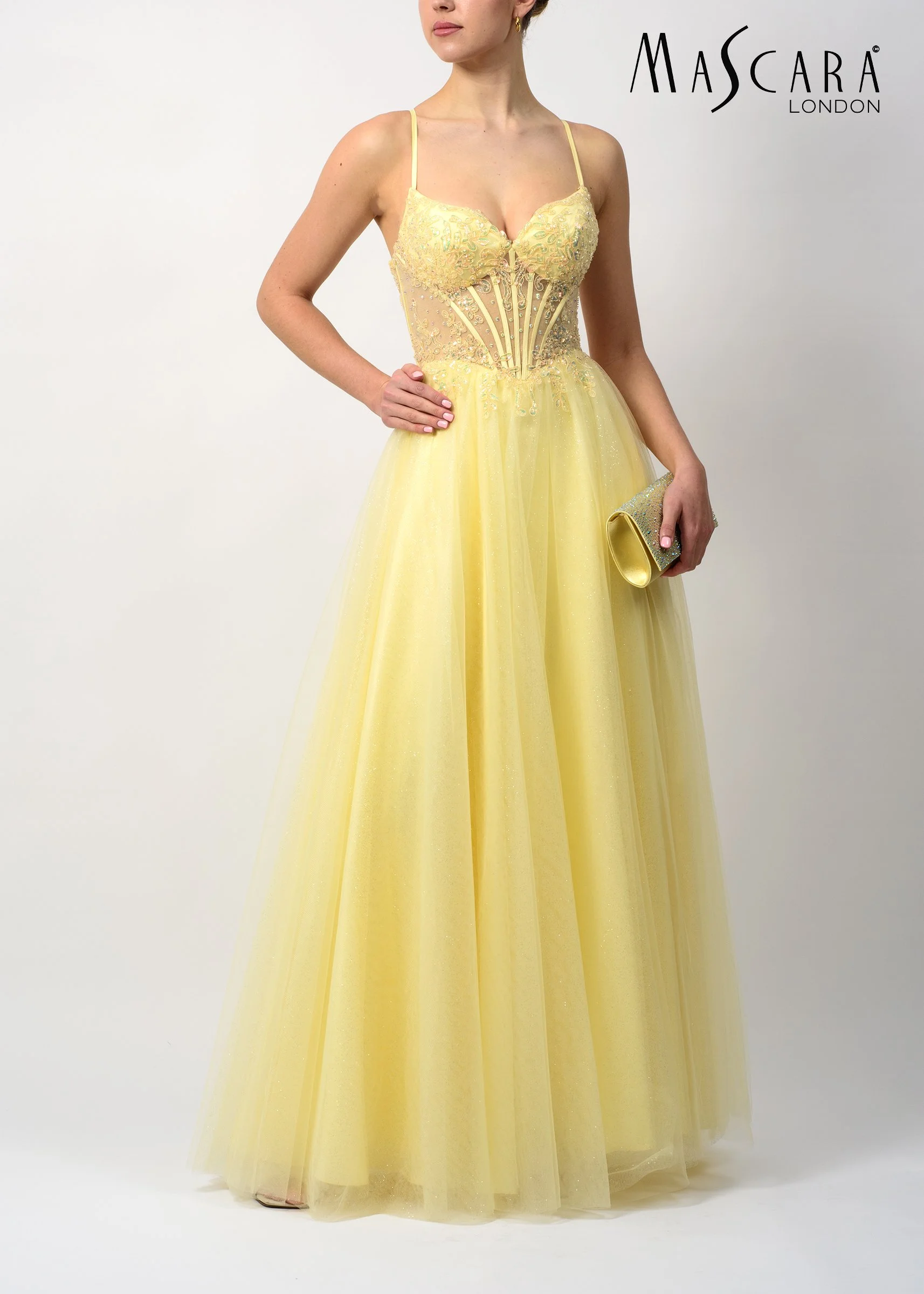 A woman in a yellow gown with intricate embroidery, holding a matching clutch, standing against a plain white background with the 'Mascara London' logo in the top right corner.