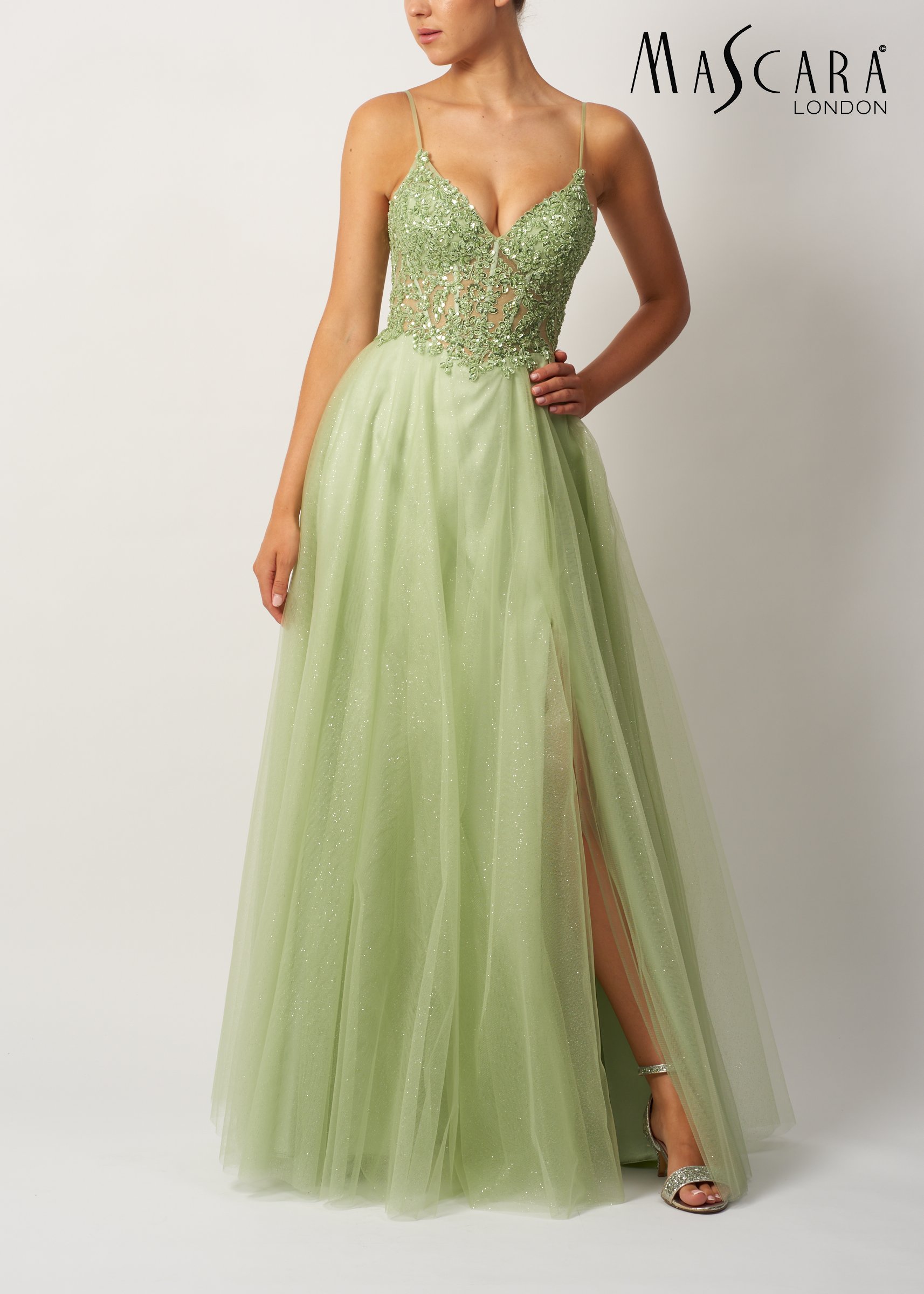 Woman wearing a lime green gown with lace and embroidery top, sheer waist, and flowing skirt, with a high slit showing her leg and silver high-heeled sandals, against a plain background, Mascara London logo in top right corner.