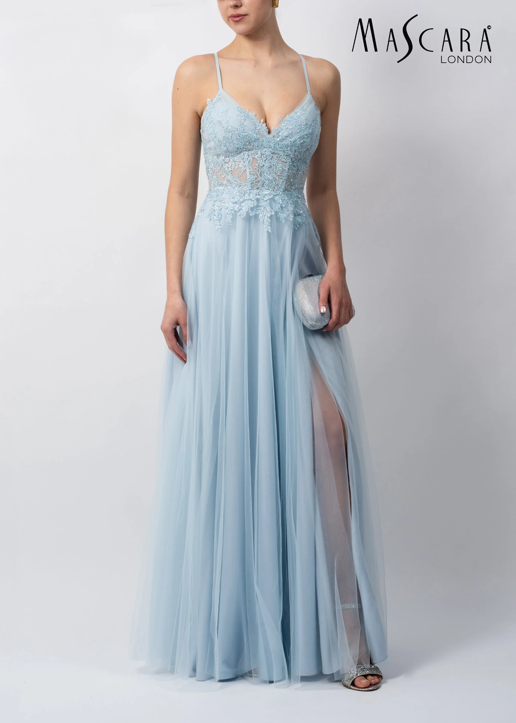 A woman wearing a light blue, lace and tulle evening gown with spaghetti straps and a slit up one leg, holding a small silver clutch, standing against a plain white background, with the text "MASCARA LONDON" in the upper right corner.