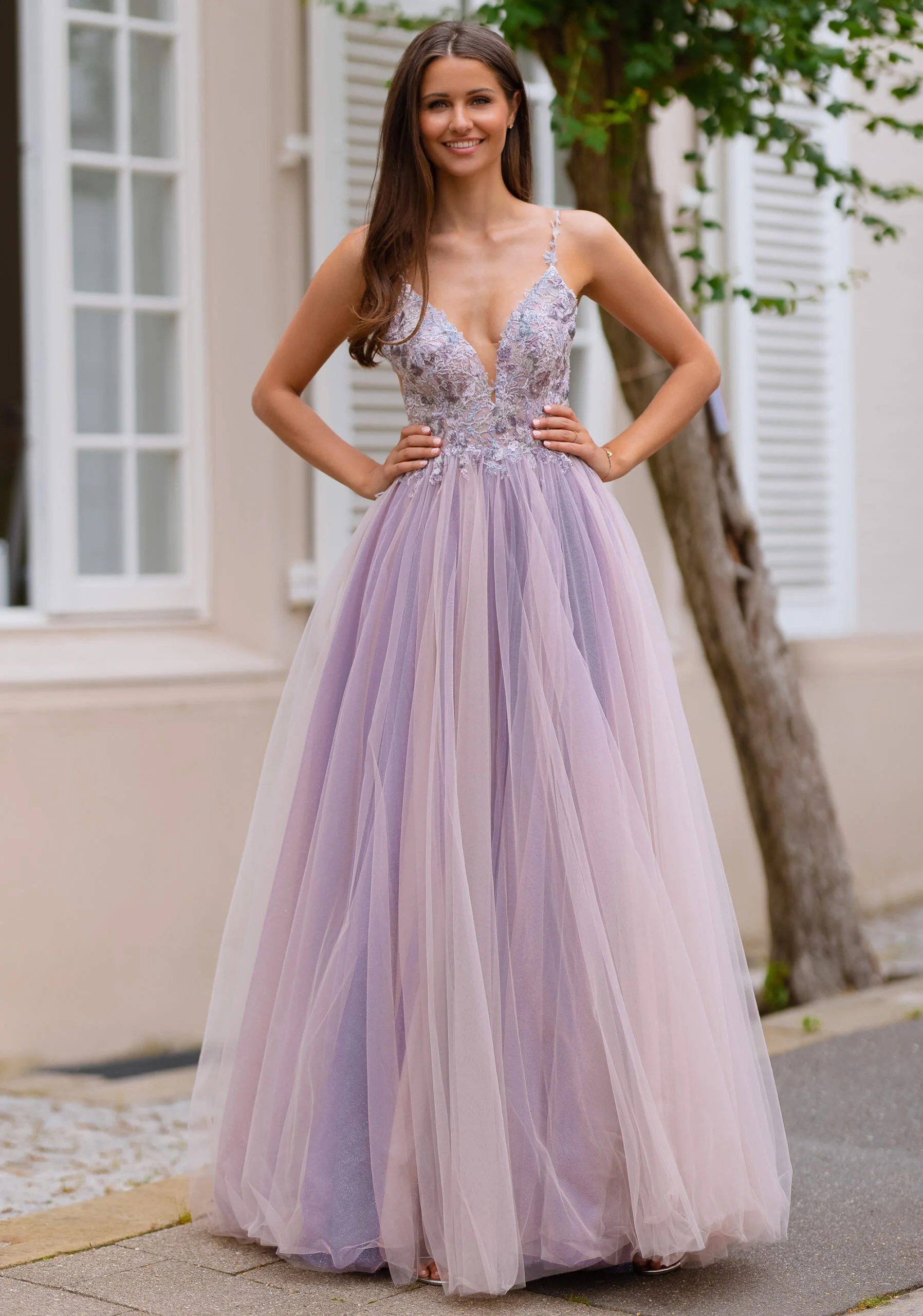 A woman in a lavender gown standing outdoors near a tree with white shutters and a window in the background.