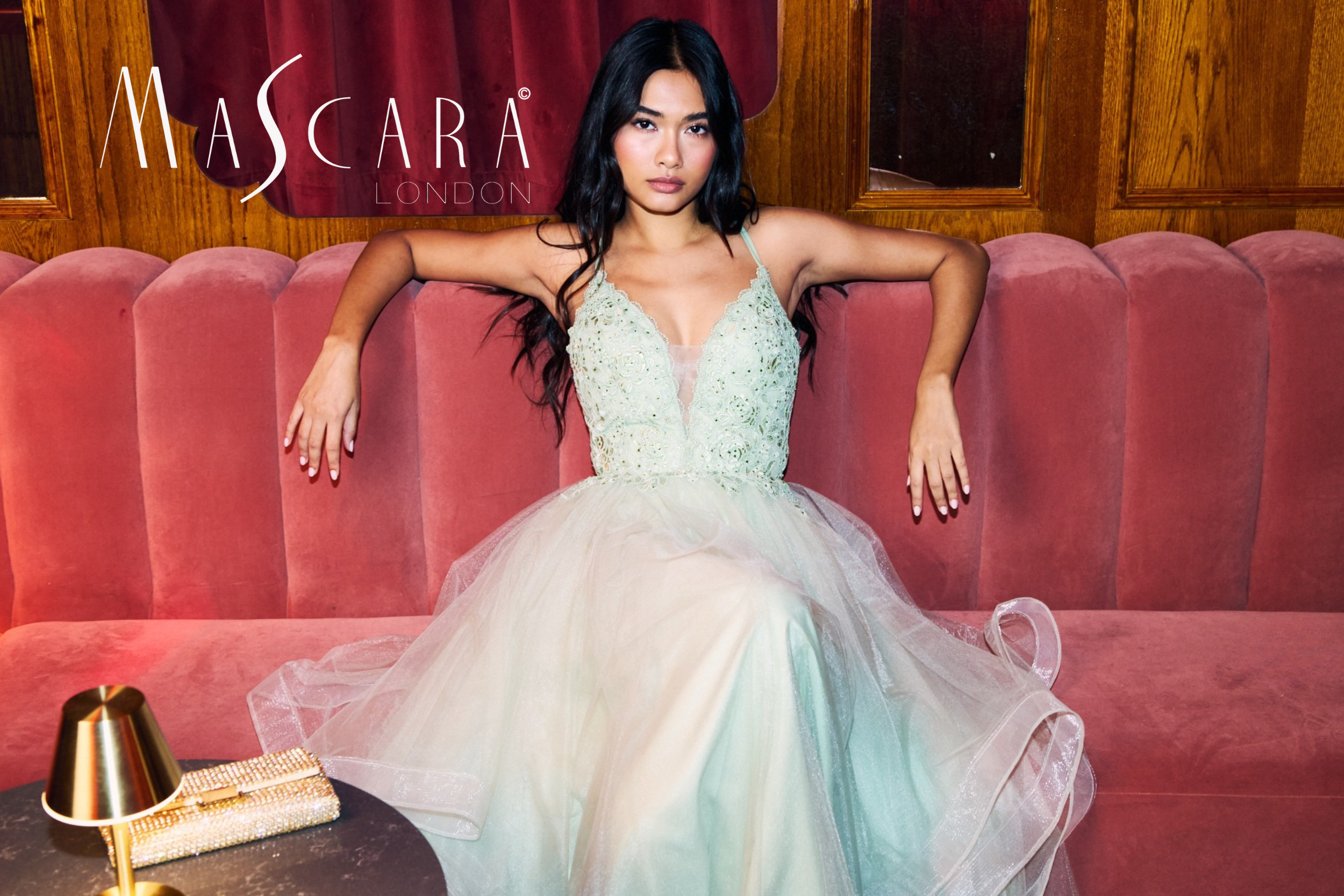 A woman in a strapless, lace and tulle dress sitting on a pink velvet couch in a room with wooden panel walls, with a gold cone-shaped jewelry stand and a gold clutch on a table in front of her.
