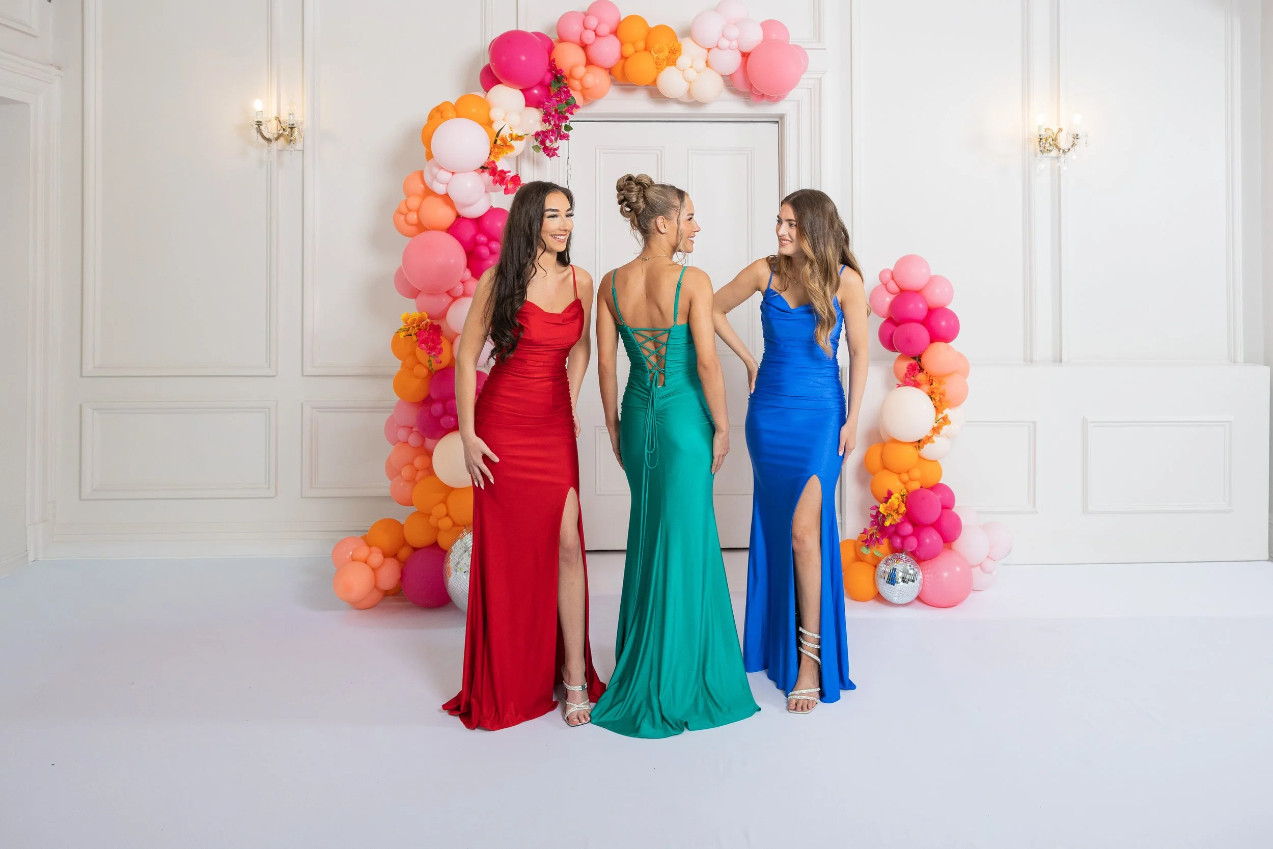 Four women in colorful evening dresses standing in front of a balloon arch at a celebration or party, smiling and engaging with each other.