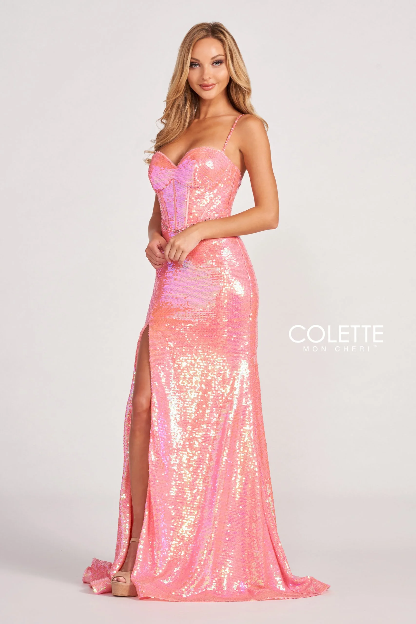 A woman wearing a long, pink, sequined gown with a high slit, standing against a plain light background. The gown has thin straps and a sweetheart neckline. She has long, wavy blonde hair and is smiling slightly.