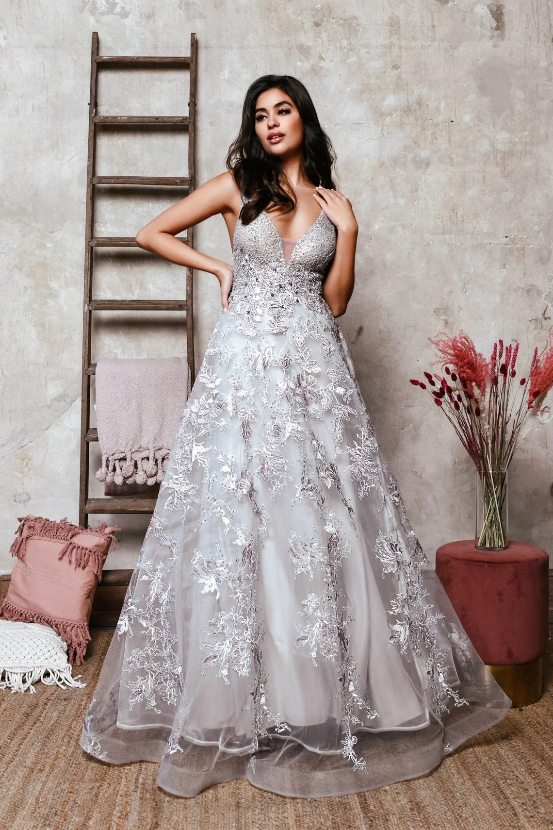 Woman in a silver and white lace prom dress standing indoors against a concrete wall, with a wooden ladder, pink pillow, blanket, and a vase of pink dried flowers nearby.