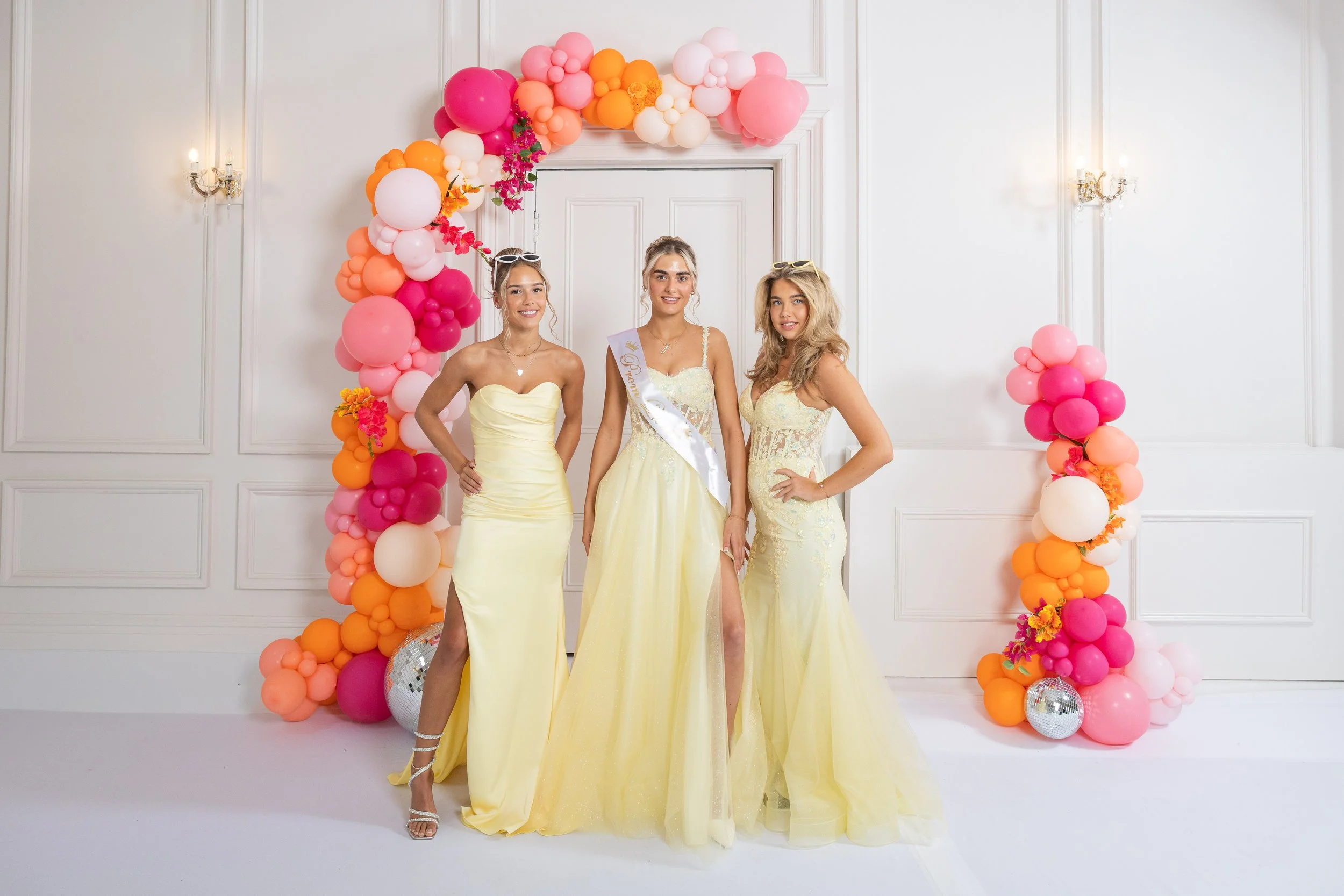 Three young women in yellow dresses posing in front of a balloon arch at a celebration, with white walls and wall sconces in the background.