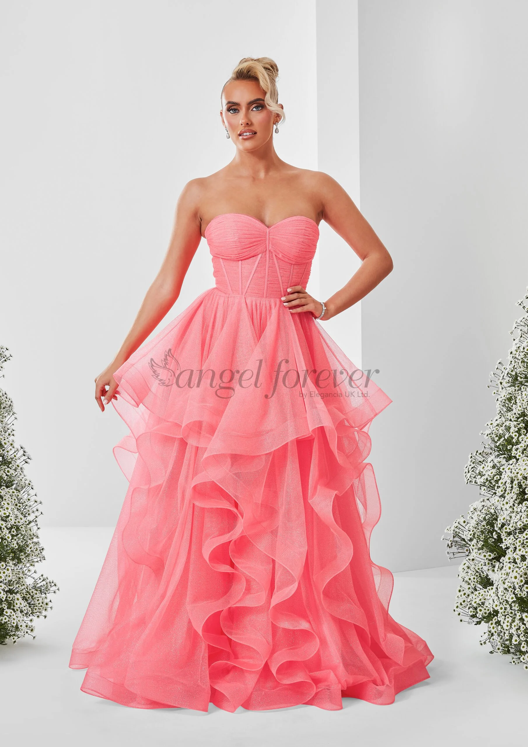 A woman wearing a strapless pink gown with a ruffled, layered tulle skirt, standing with one hand on her hip in a neutral background with floral arrangements on each side.