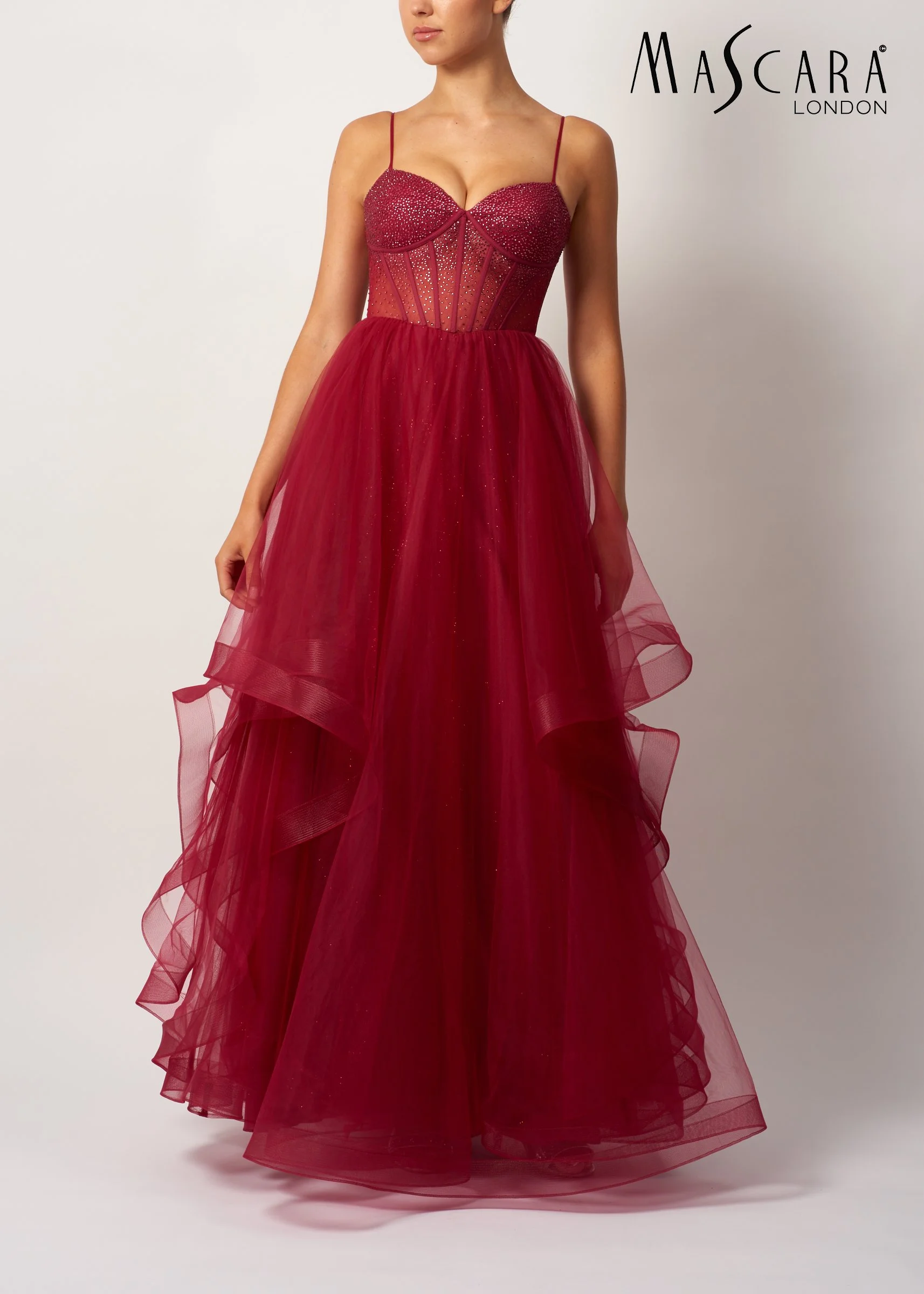 A woman wearing a red, strapless, embellishment dress with a fitted bodice and flowy layers of tulle, standing against a plain white background with the text 'Mascara London' in the upper right corner.