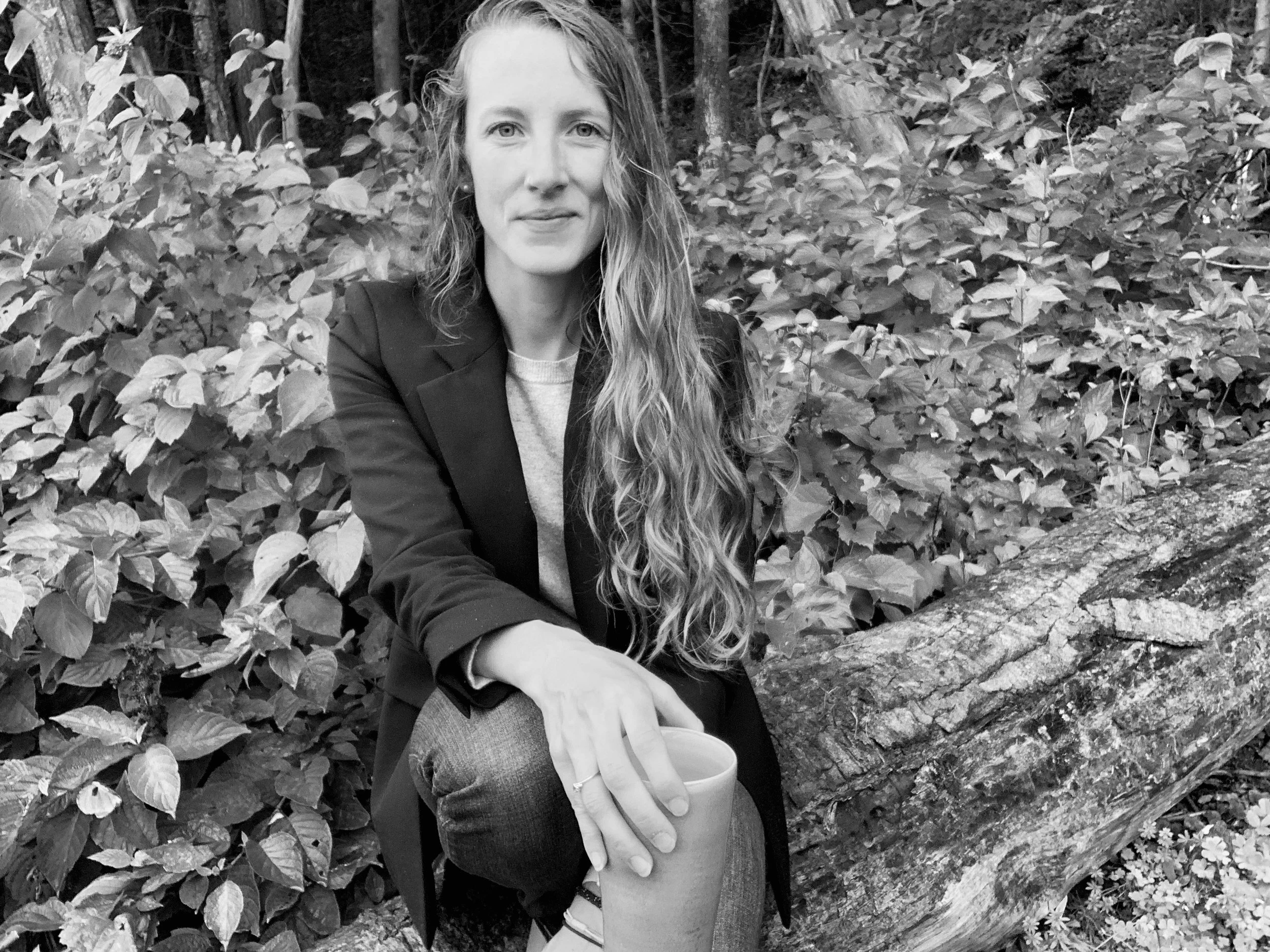 A woman sitting outdoors in front of dense foliage, holding a cup with both hands, smiling slightly, dressed in a blazer and casual clothing.
