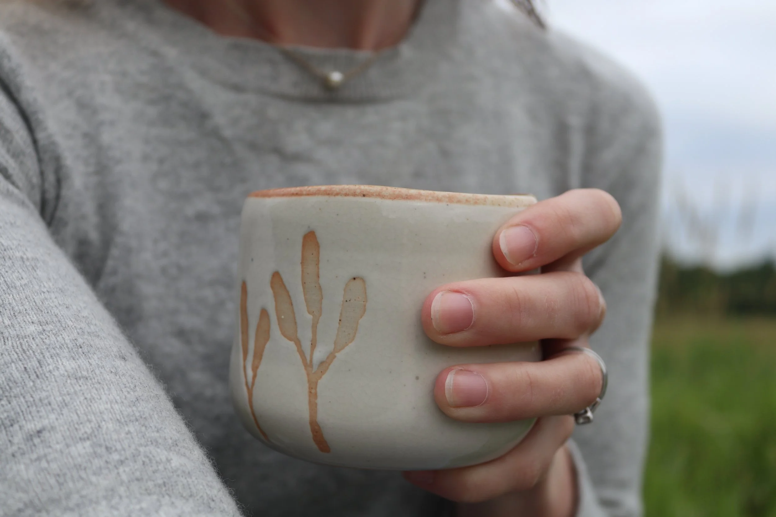 Person holding a ceramic mug with a hand-painted tree design, wearing a gray sweater and silver ring, with a blurred outdoor background.