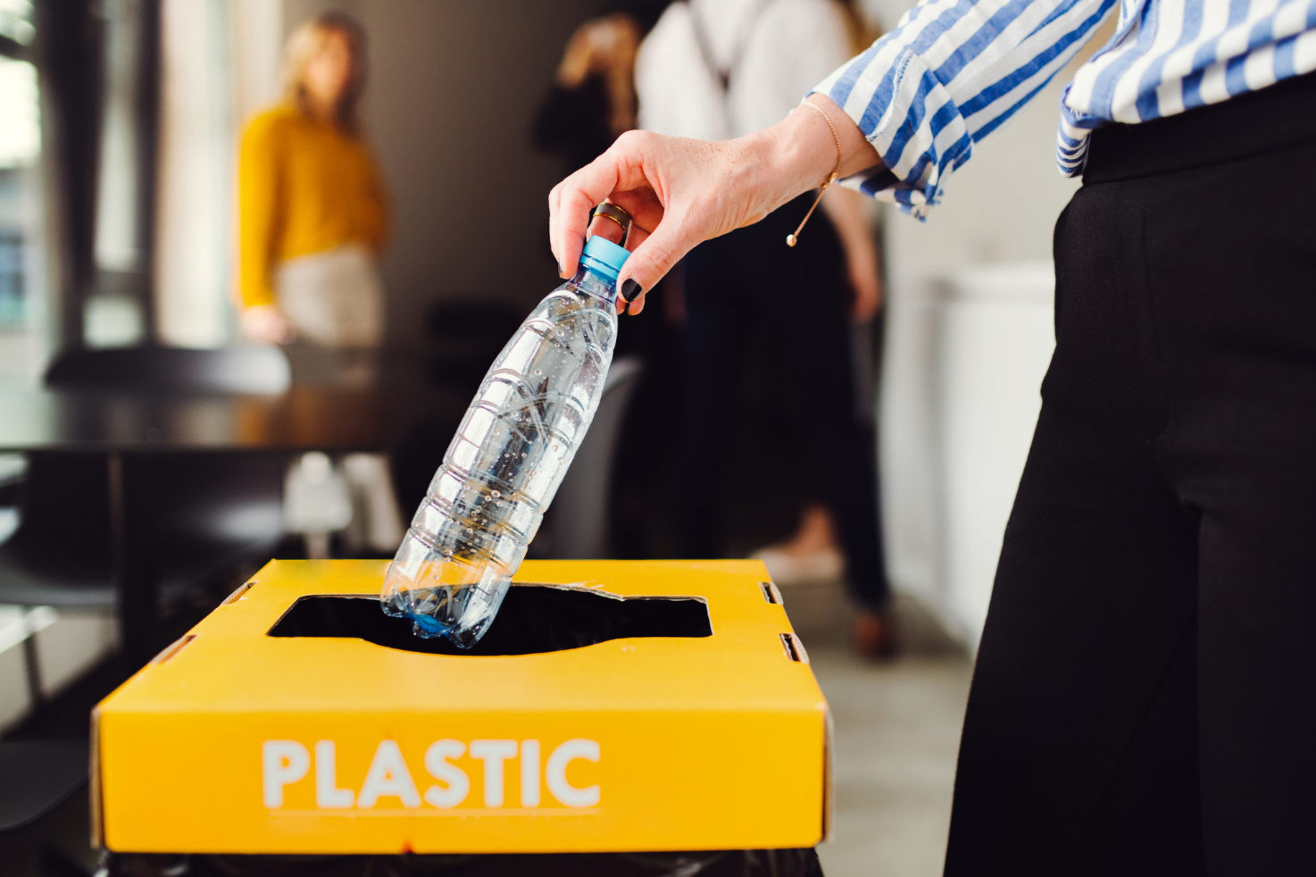 A person discards a plastic water bottle into a yellow recycling bin labeled 'PLASTIC' in an office or conference room setting.