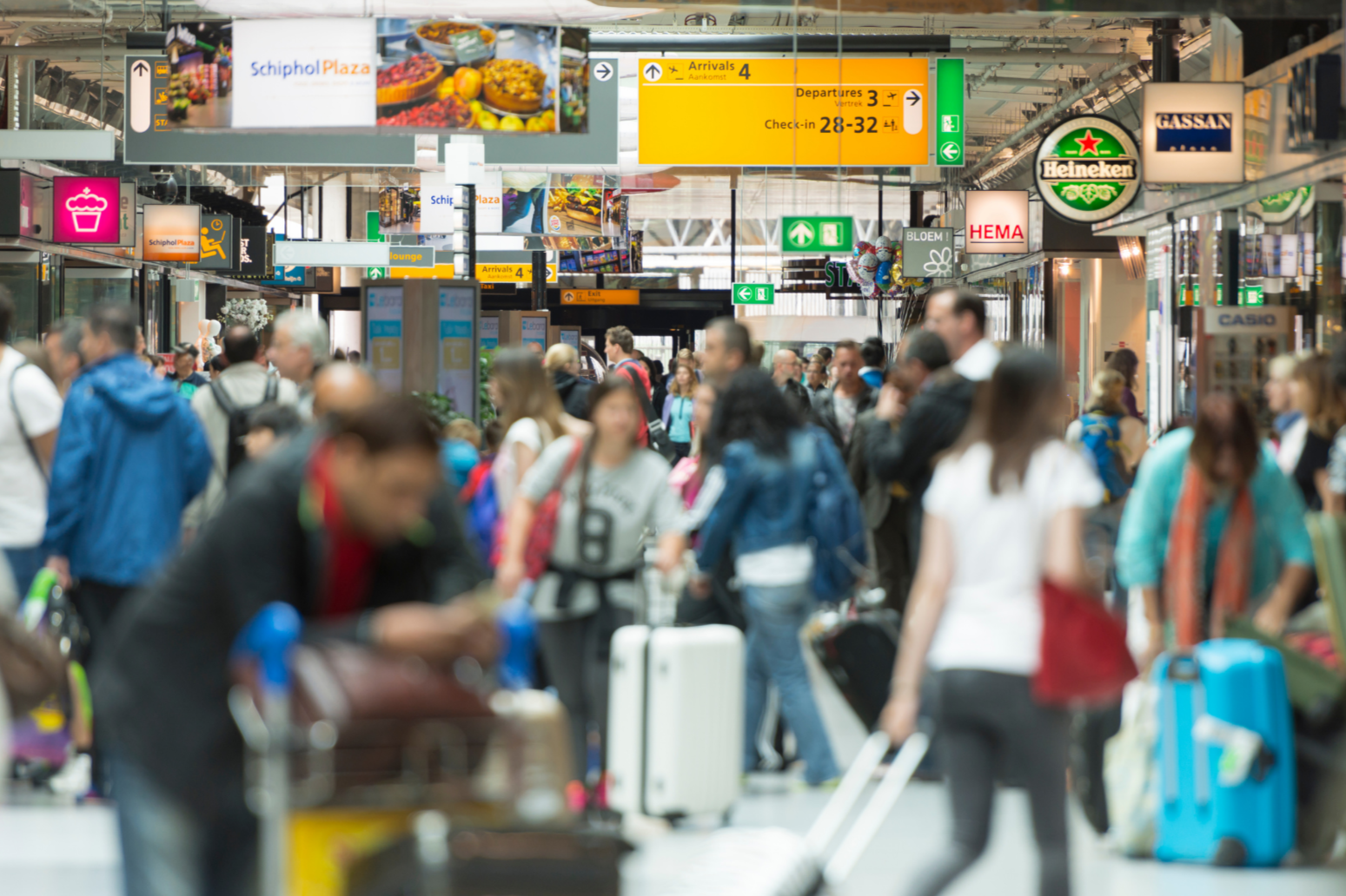 Crowd of travelers at an airport terminal with signs for arrivals, departures, check-in, and shops such as Heineken and Gassan.