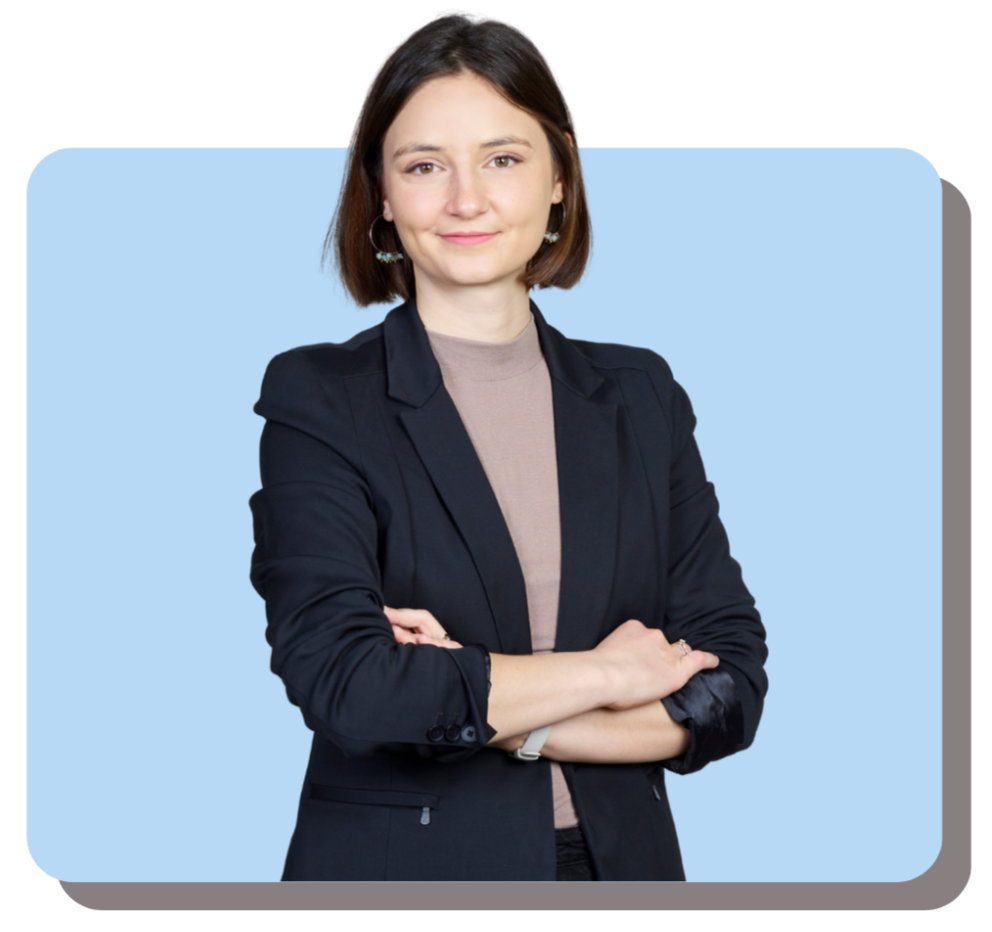 A woman with short brown hair, wearing a black blazer and a beige shirt, standing with arms crossed and smiling against a light blue background.