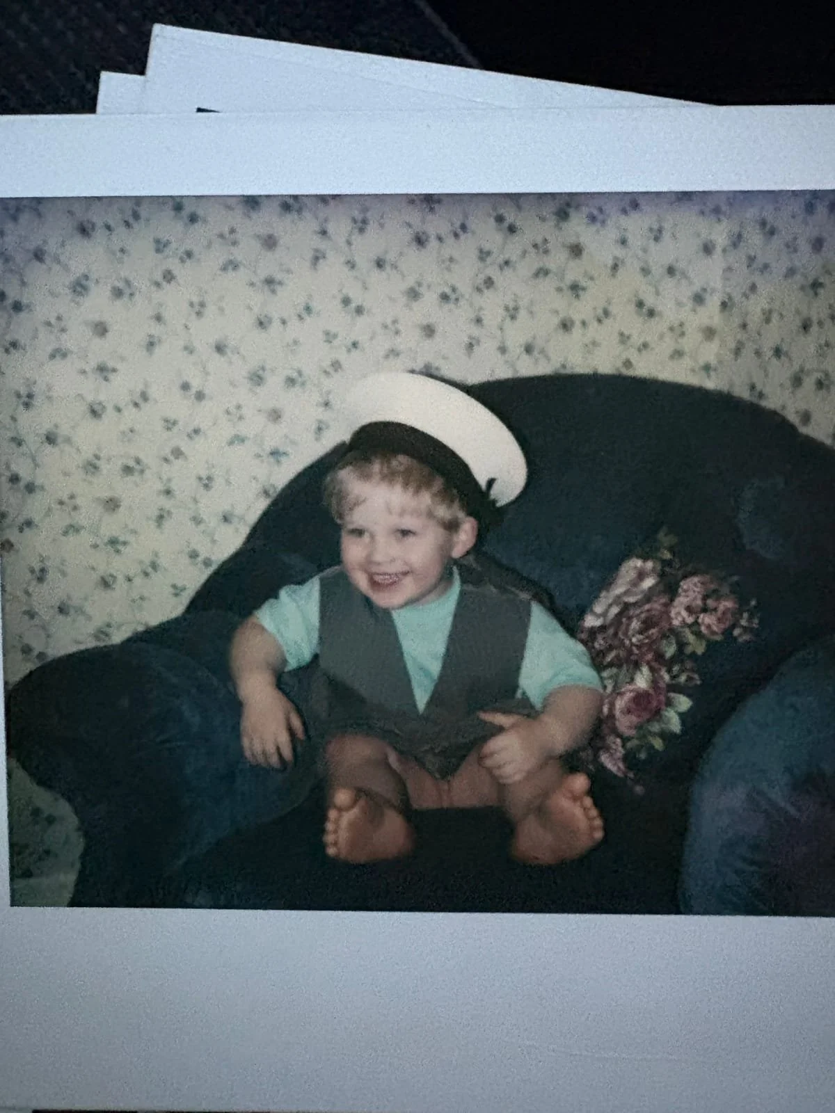 Young child smiling while wearing a sailor hat and waistcoat during imaginative play at home.
