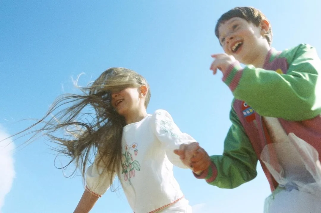 Two children, a girl with long hair and a boy, holding hands and laughing outdoors under a clear blue sky.