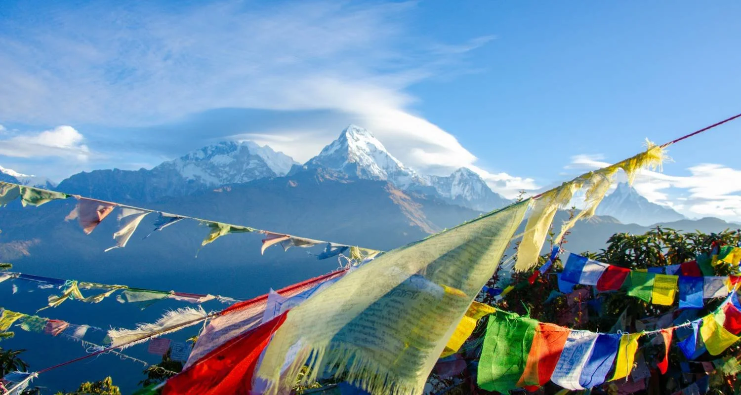Colorful prayer flags flutter in front of a mountain range with snow-capped peaks under a blue sky.