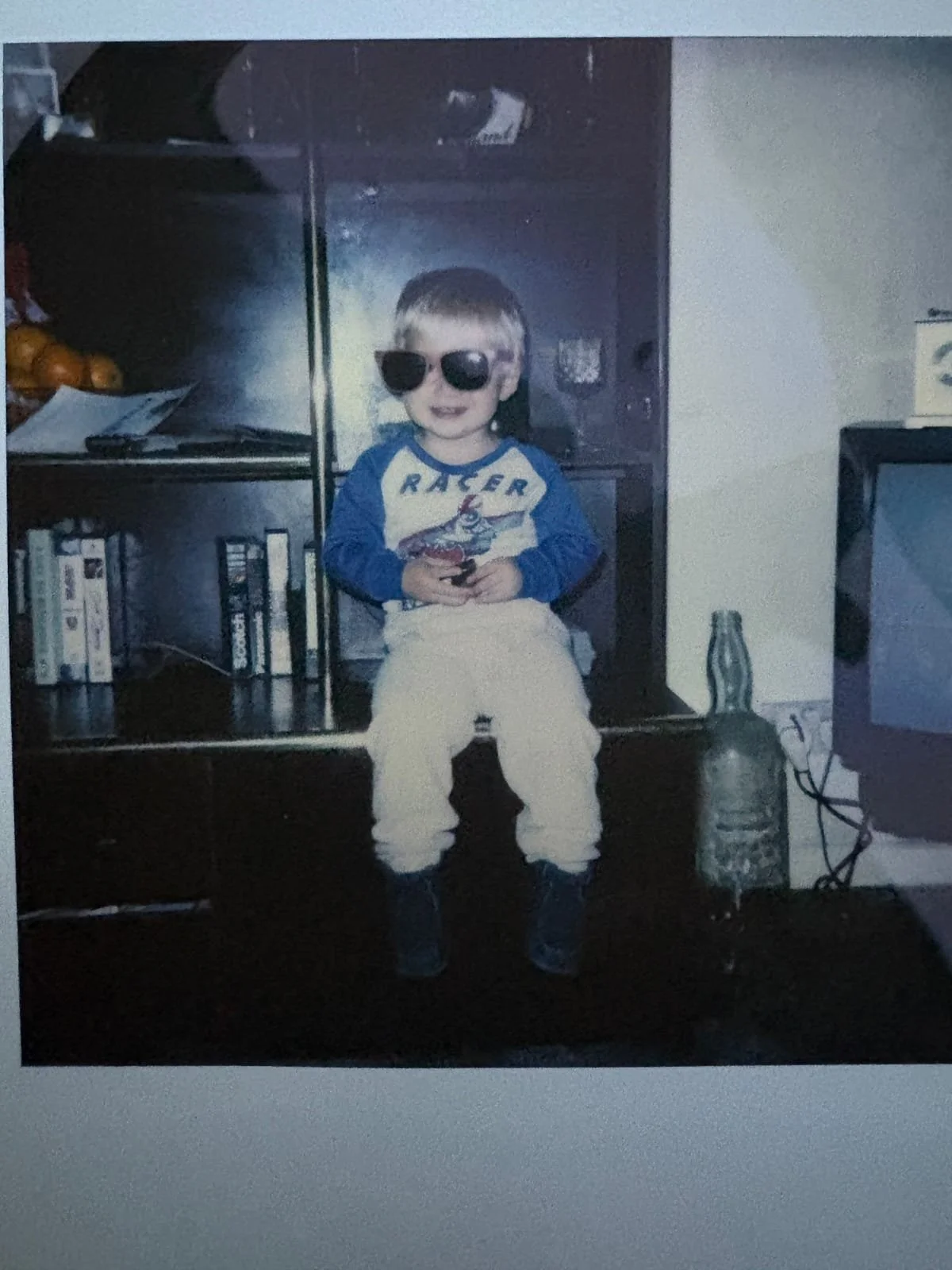 Young child wearing oversized sunglasses while sitting on a television cabinet at home.