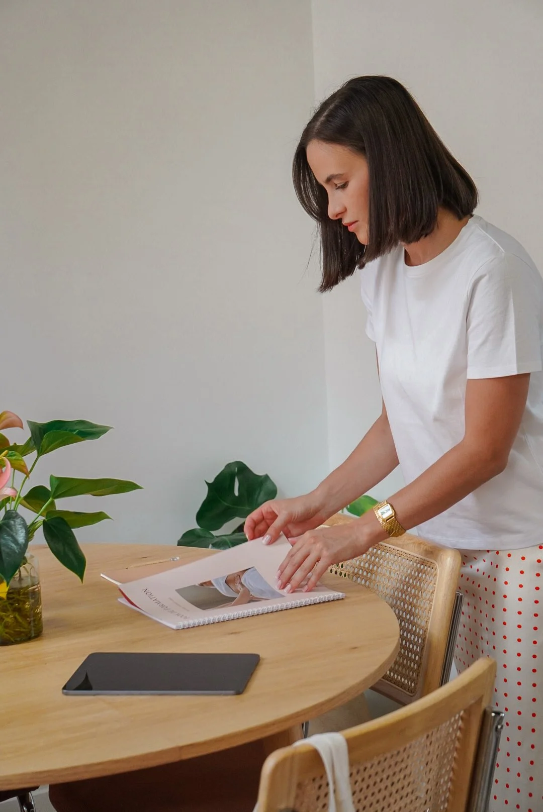 Femme debout à côté d'une table en train de tourner les pages d'un cahier. Il y a des plantes vertes à gauche, une tablette et une tasse en verre sur la table.