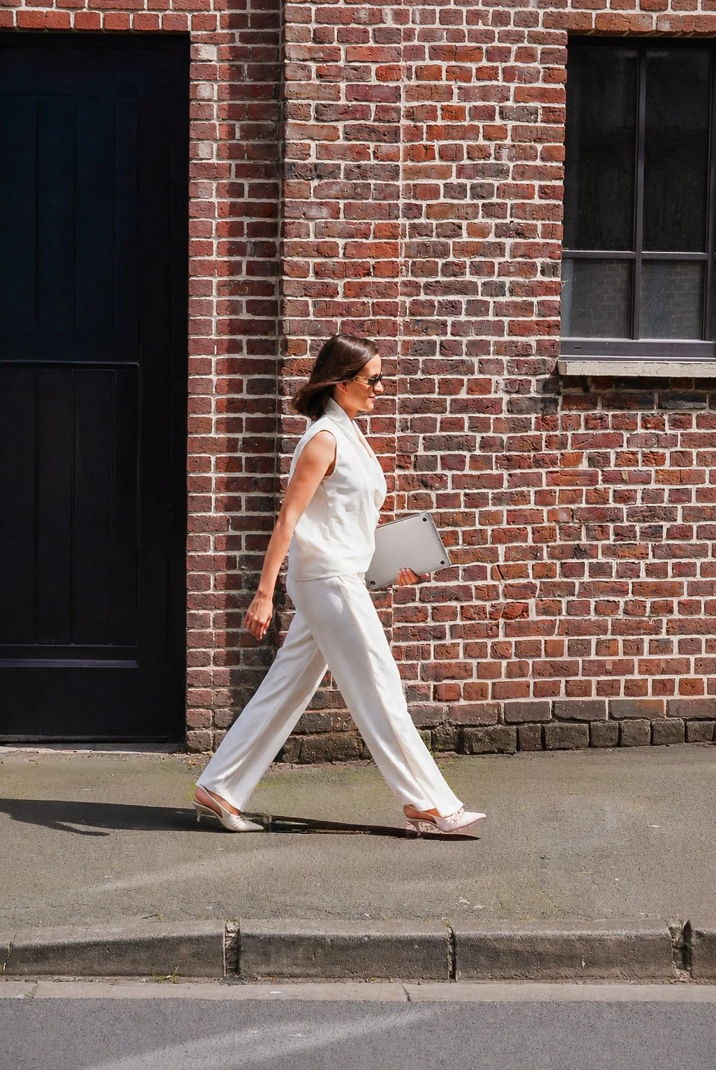 Femme en tenue blanche marchant sur le trottoir près d'un mur en briques, tenant un ordinateur portable.