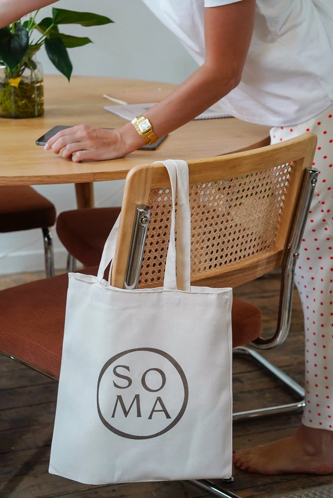 Une personne debout près d'une table en bois, avec un sac en toile blanc suspendu à la chaise en bois à l'avant-plan, portant un logo avec le texte 'SOMA'. La personne porte un bracelet doré et des vêtements blanche avec des points rouges.
