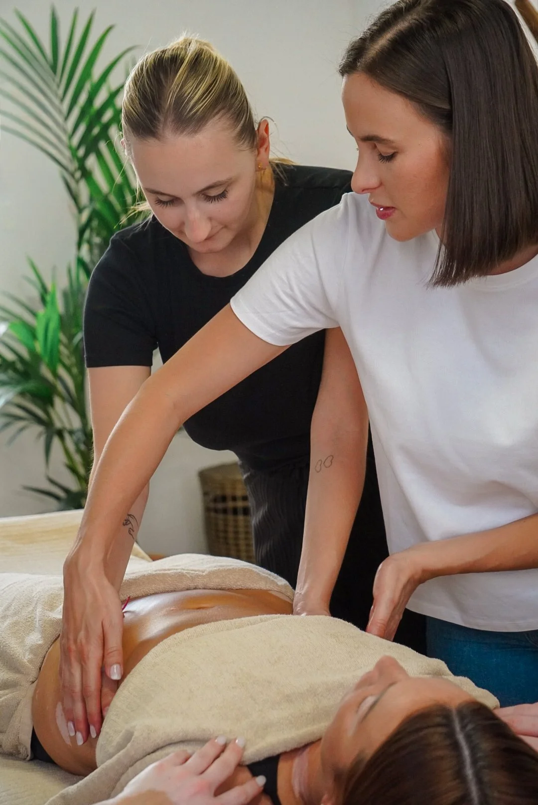 Deux femmes pratiquent une séance de massage ou d'acupuncture sur une femme allongée sur une table, dans une pièce avec une plante verte en arrière-plan.
