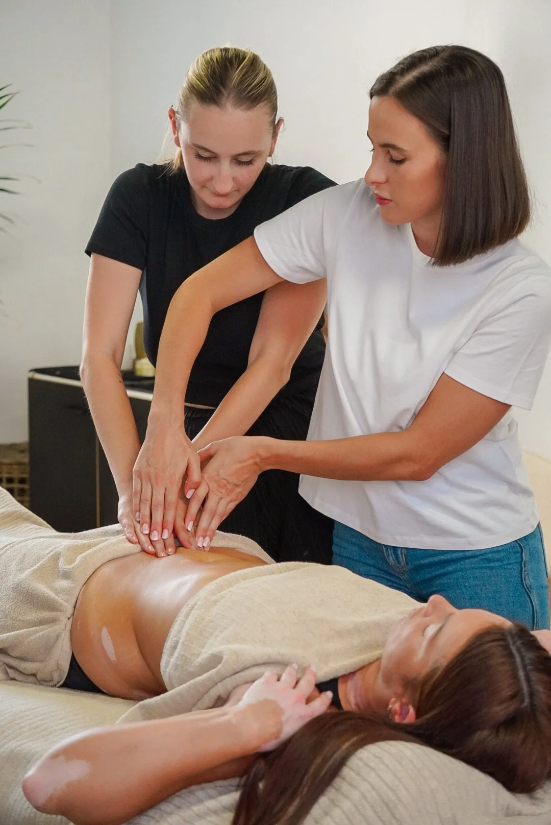 Deux femmes pratiquent un massage thérapeutique sur une femme allongée sur un lit, dans une pièce blanche.