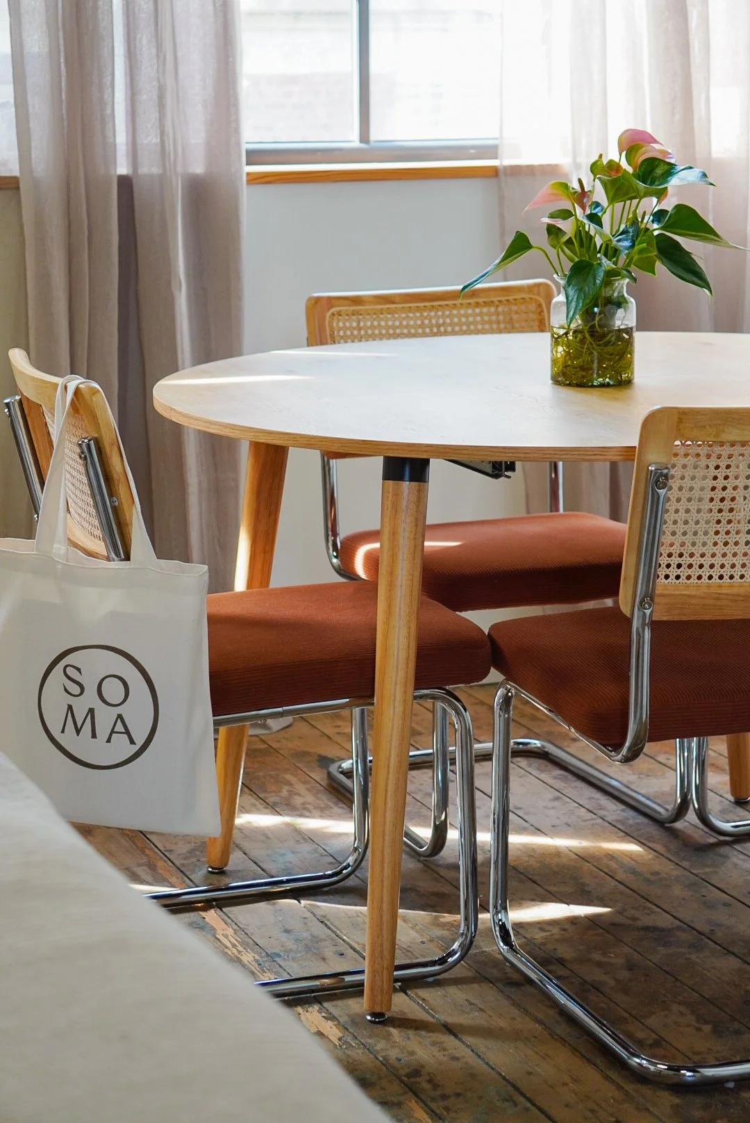 Table en bois rond avec une plante verte dans un vase en verre sur une fenêtre en arrière-plan, chaises en bois avec coussins marron, sac en toile blanche suspendu à une des chaises.