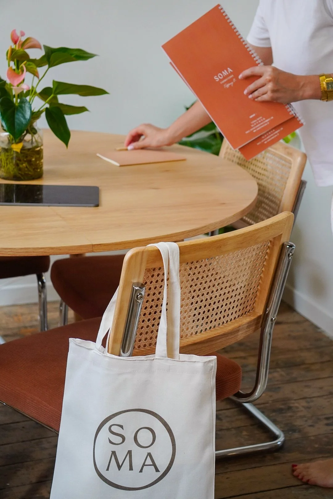 Une salle de réunion ou de travail avec une table en bois, des chaises en bois avec assises marron, une plante decorative, une personne tenant un carnet orange avec le texte 'SOMA', une sac en toile blanche avec le logo 'SOMA' suspendue à une chaise.