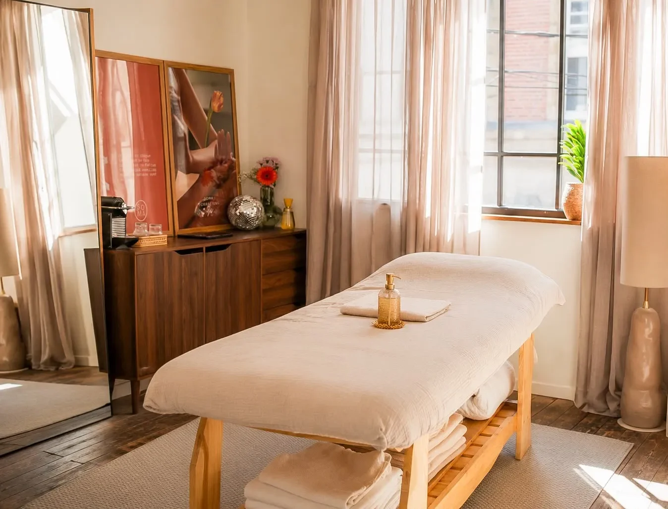 Une salle de massage avec un lit recouvert d'une draperie blanche, des serviettes pliées en dessous, et un flacon de lotion sur le lit. La pièce est lumineuse avec des rideaux pastel, une plante en pot, et un meuble en bois avec des décorations.