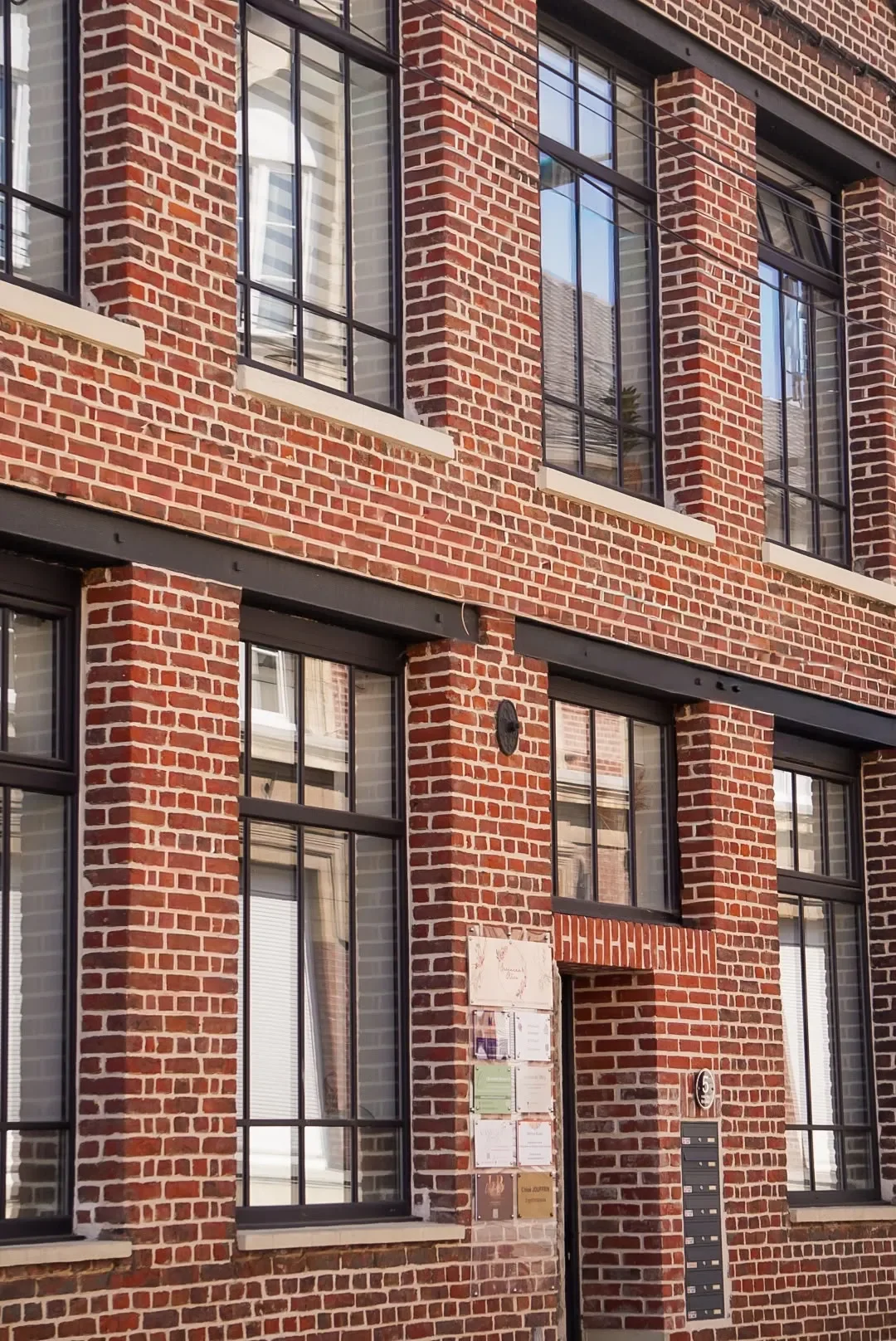 Façade d'un bâtiment en briques rouges avec plusieurs fenêtres à cadre noir, une plaque d'information et une boîte aux lettres.