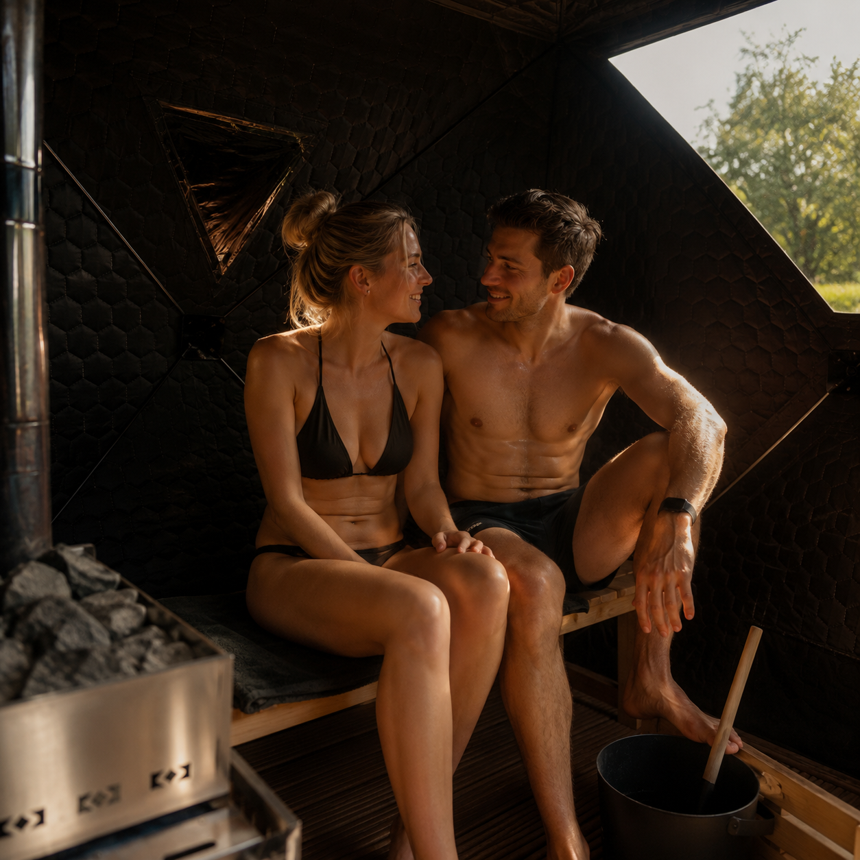 A man and woman sitting close together inside a sauna, smiling and looking at each other, with a bucket and sauna ladle nearby.