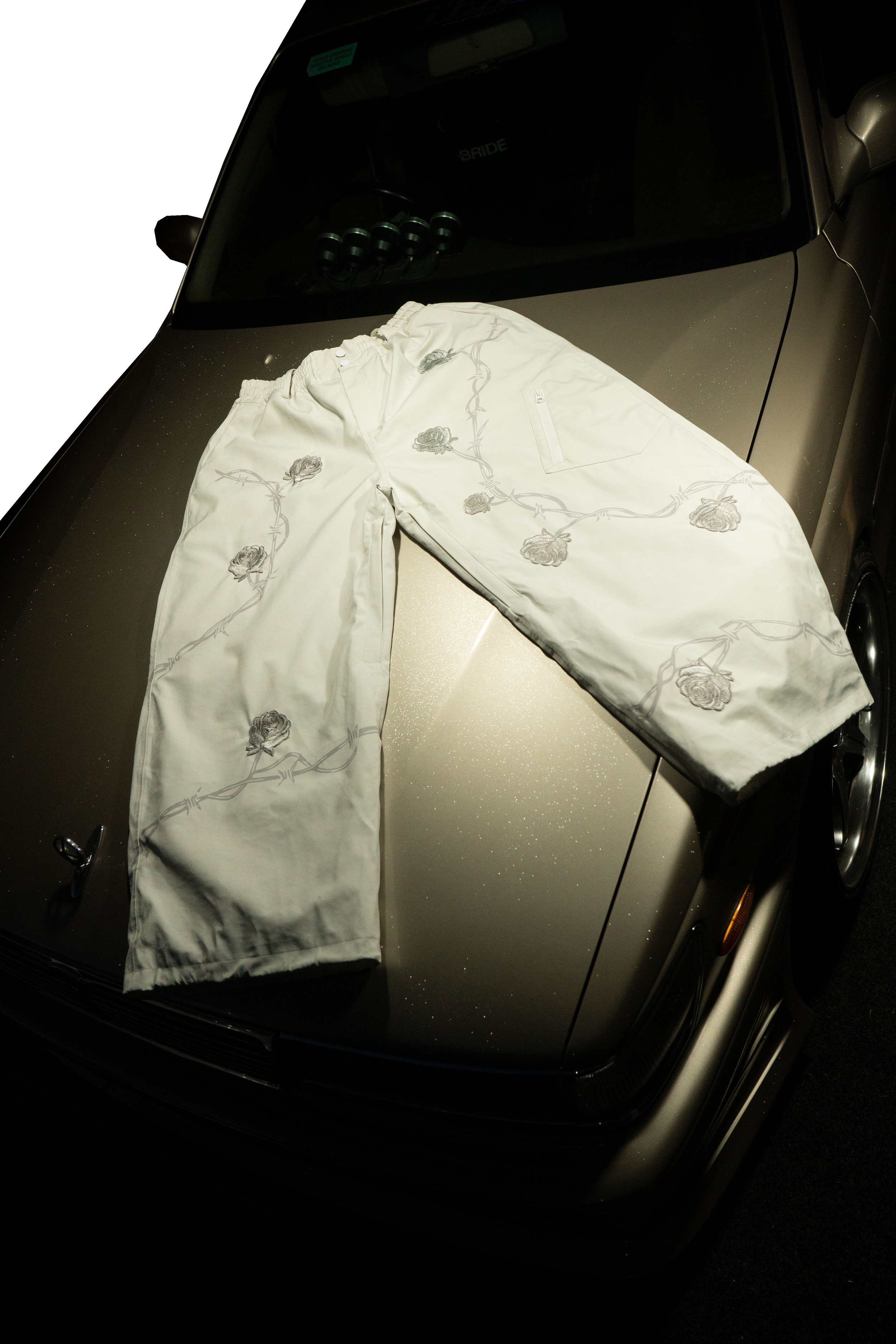 A pair of white pants with floral embroidery laid across the hood of a dark-colored car. The car is parked at night, with visible parts of the windshield, side mirror, and front wheel.