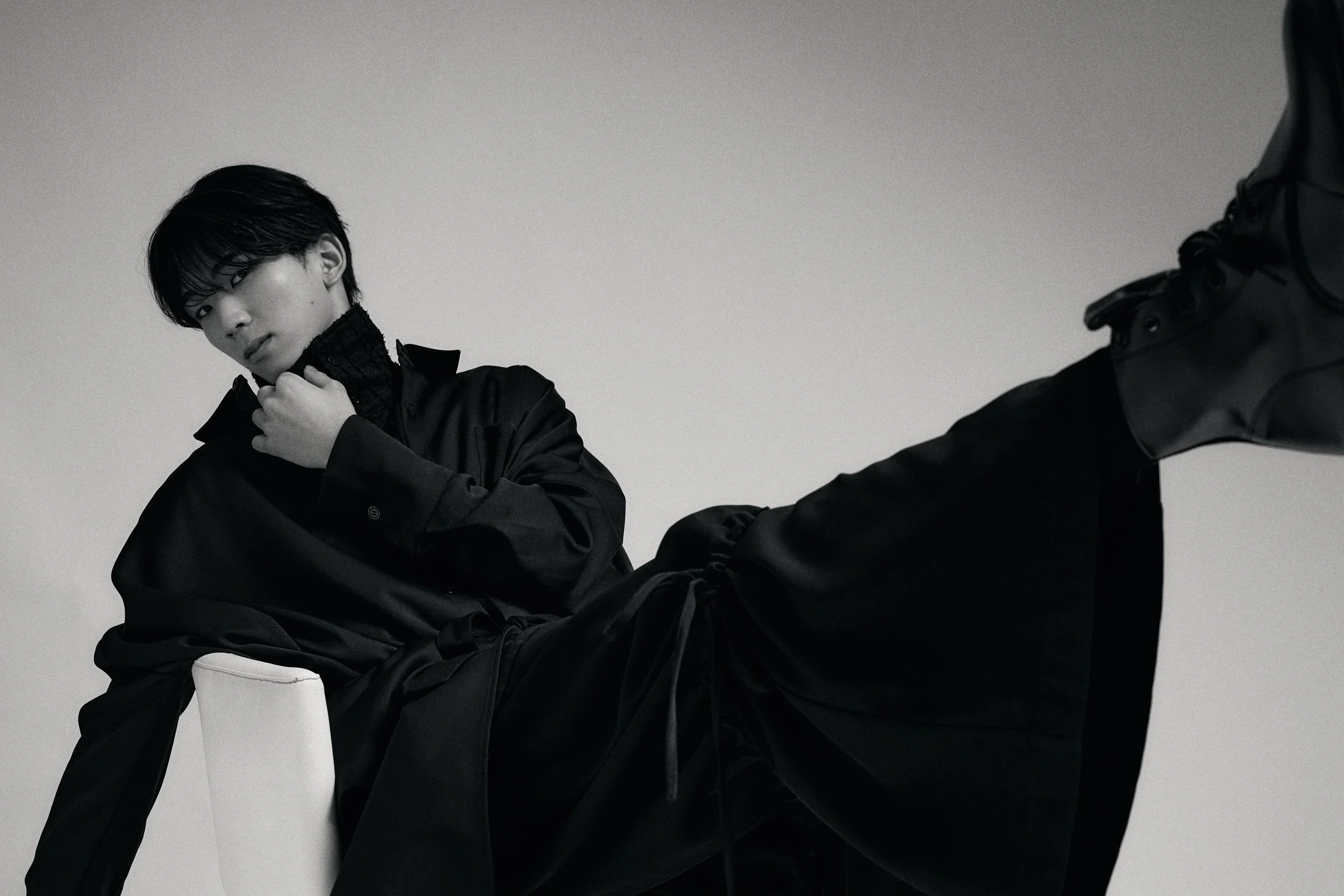 黒い服を着た若い男性が、片膝をついて座っている写真。男性は黒いジャケットとハイネックのトップスを着ており、黒髪で髪は少し濡れている様子。背景はシンプルなグレー。