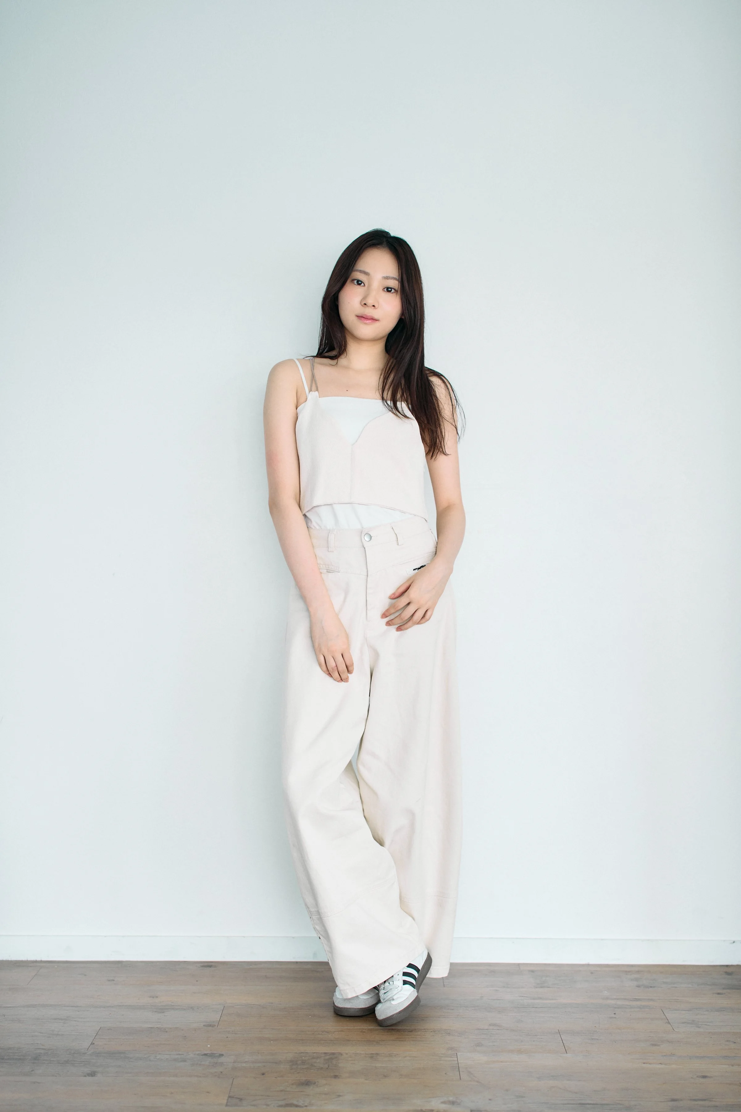 白い壁の前に立つ若い女性。白いタンクトップとゆったりとした白いパンツを穿き、スニーカーを履いている。