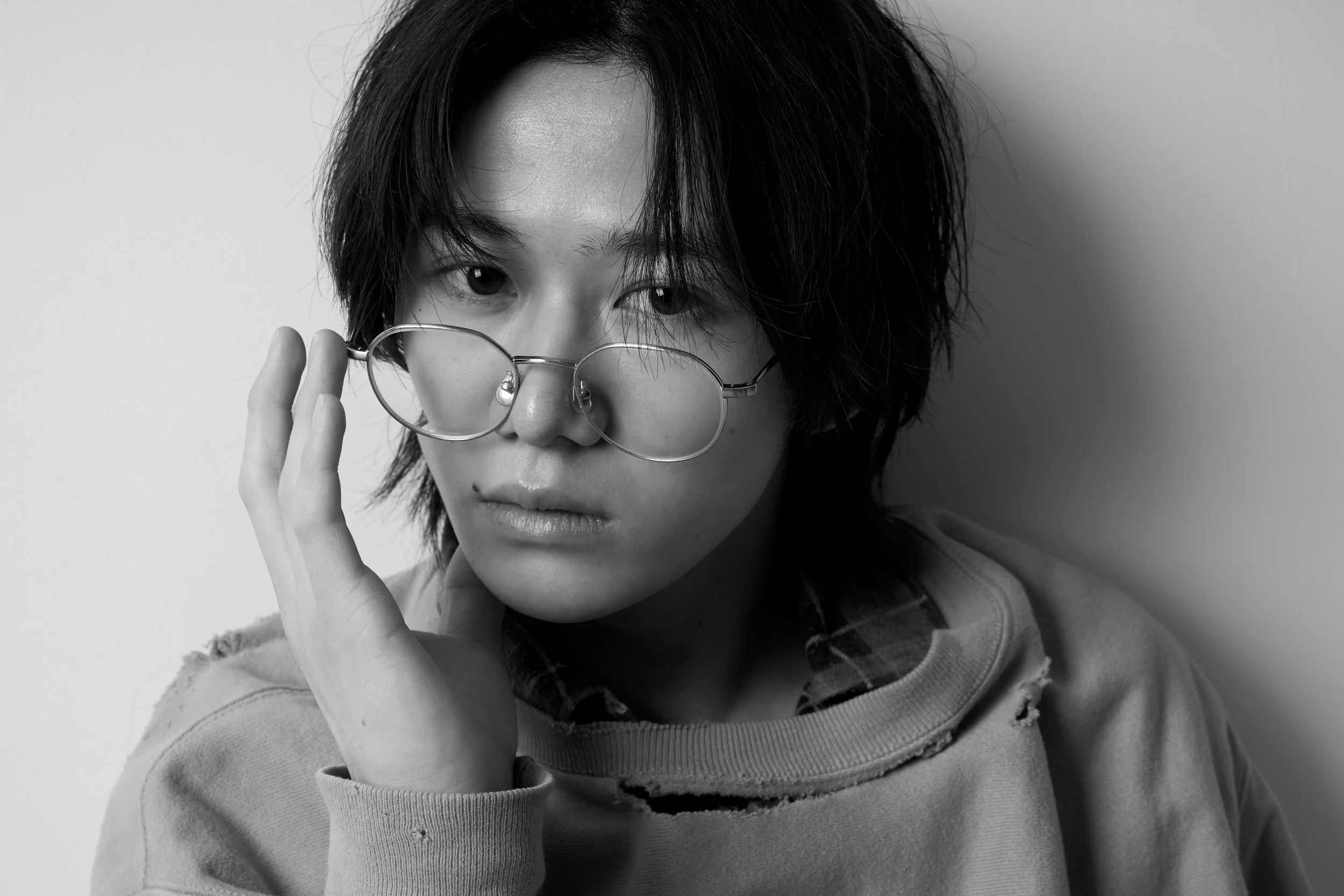 黒髪の若い男性が丸眼鏡を持ち上げながらじっと見つめている黒と白の写真