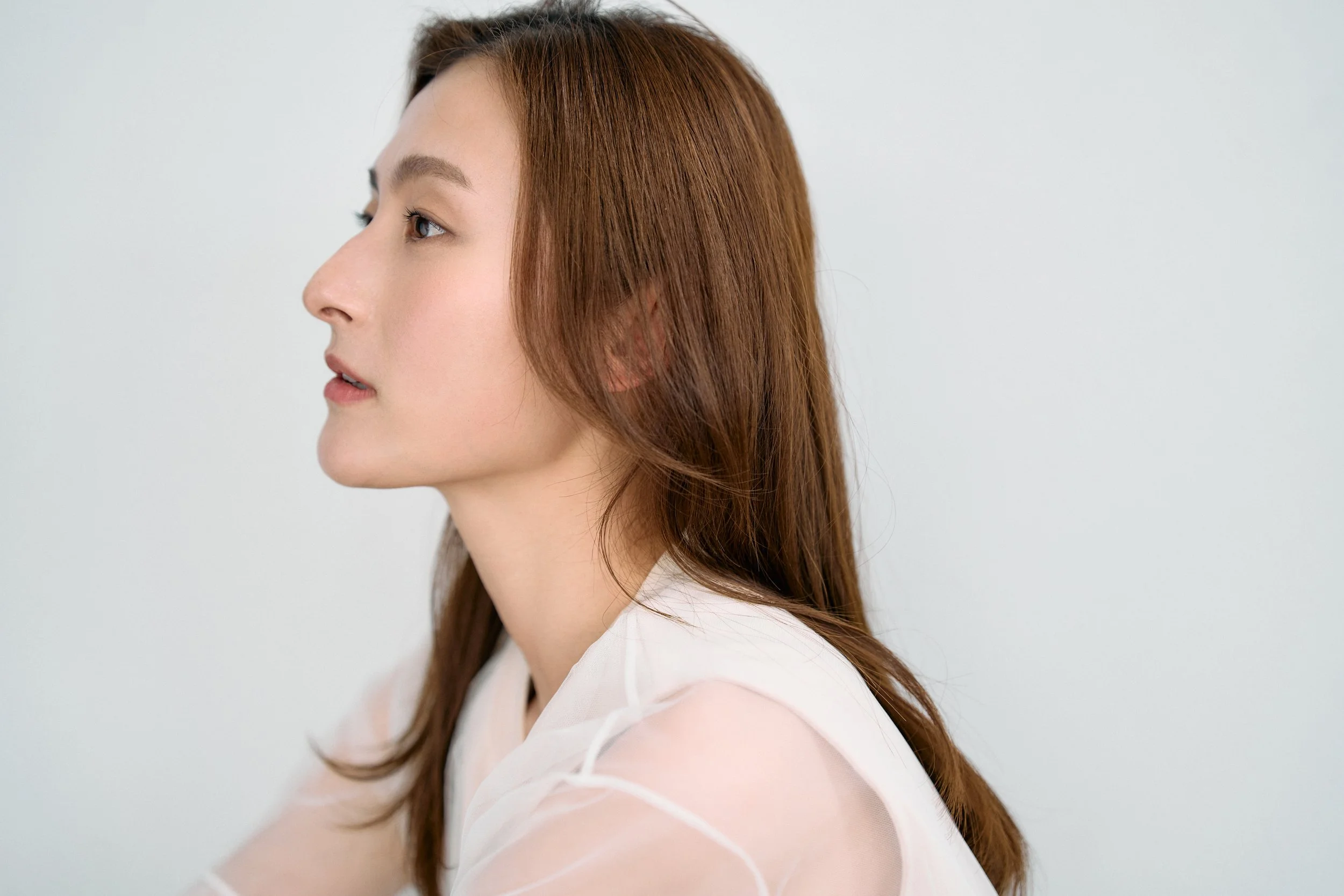 側面から見た若い女性の顔と肩、茶色の長い髪、白い衣服、淡い背景。