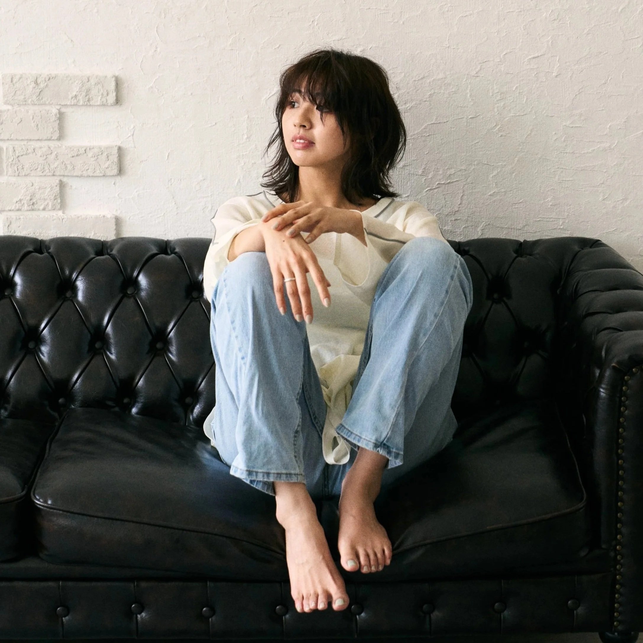 白い壁の前に座る若い女性がカジュアルな服装で座っている。彼女は肩までの濃い茶色の髪を持ち、リラックスした表情をしており、左手を胸に置き、右手は膝に置いている。彼女は黒いレザーカウチに座っている。